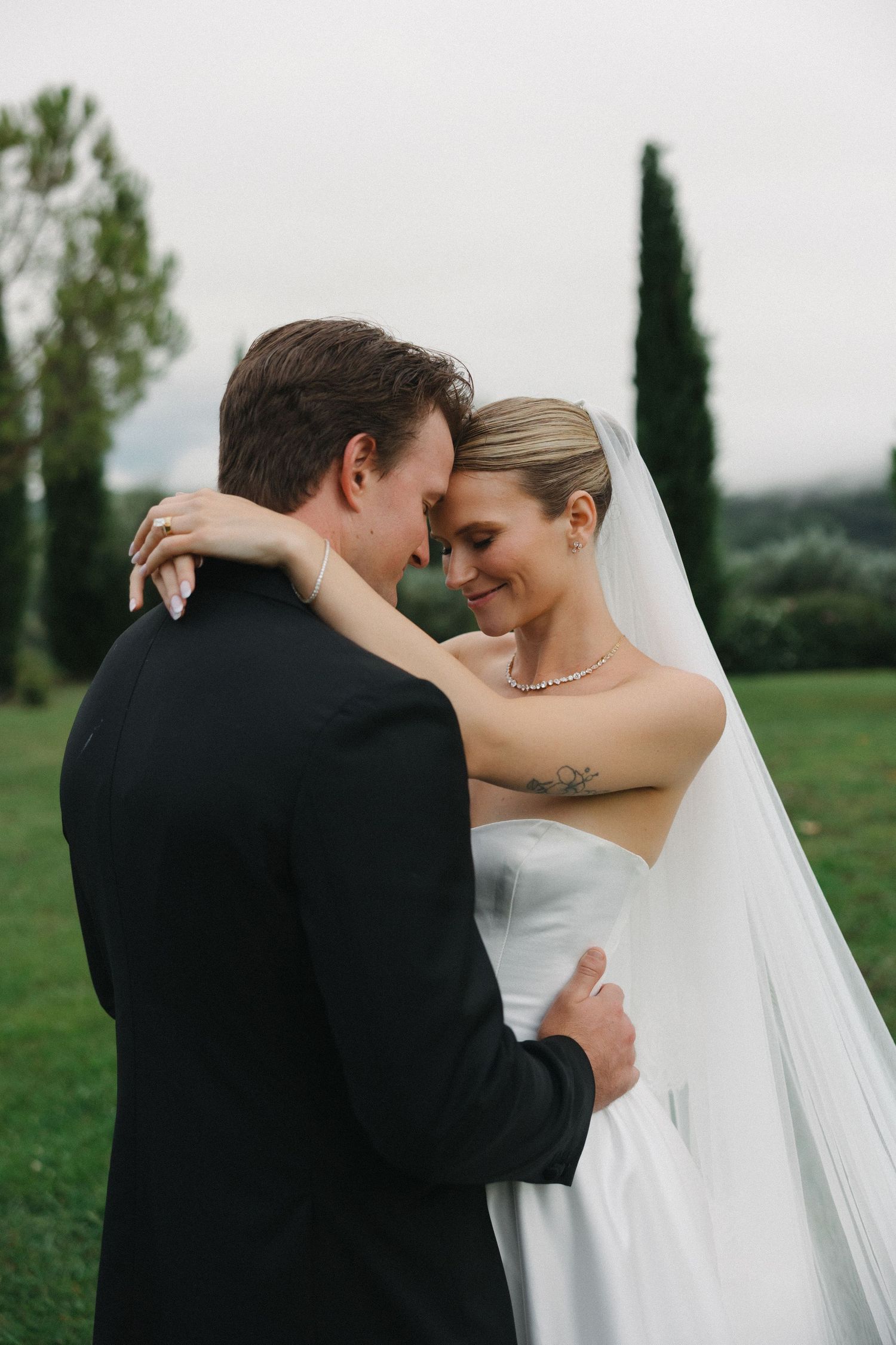 Bride and groom share an intimate moment outdoors, touching foreheads, with cypress trees and green hills in the background.