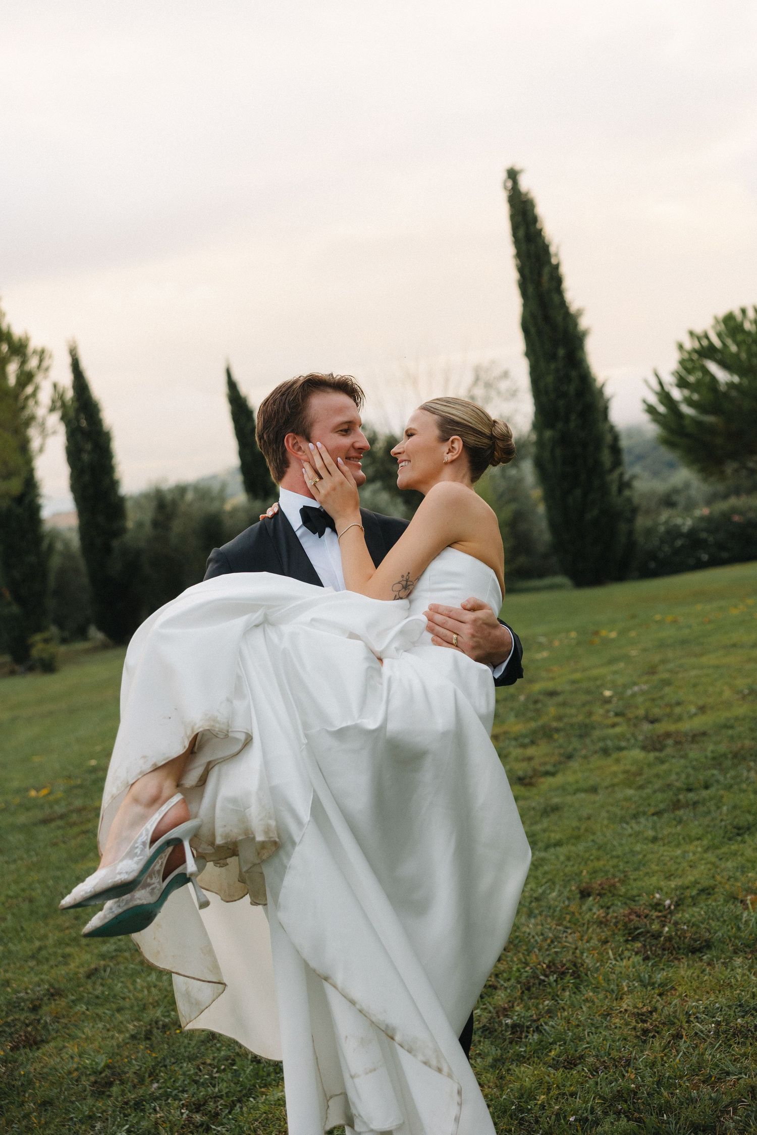 Groom carrying bride in white gown outdoors, sharing a romantic moment with cypress trees in background.