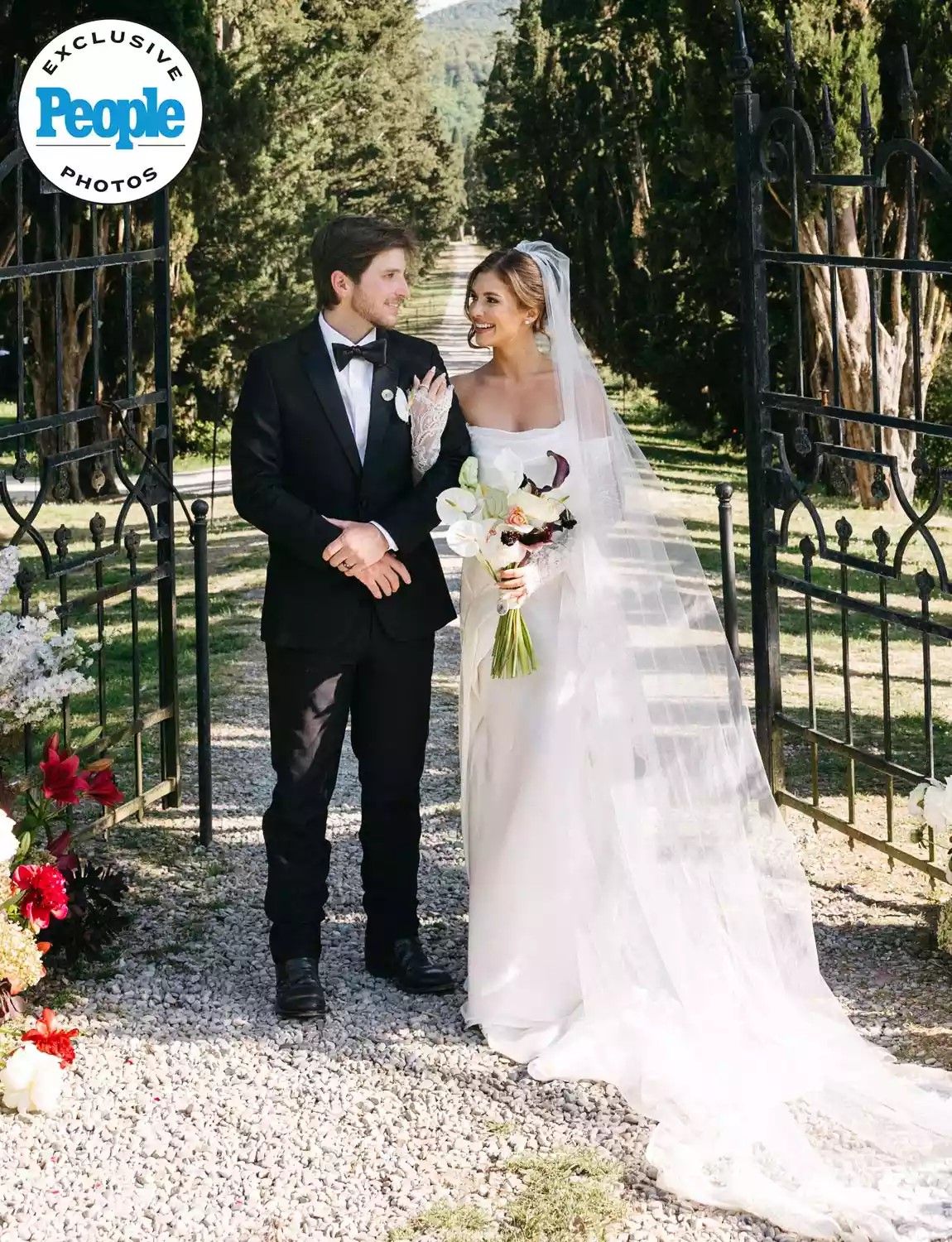 Couple in wedding attire smiling at iron gate, bride in white gown with veil holding bouquet, groom in black tuxedo.