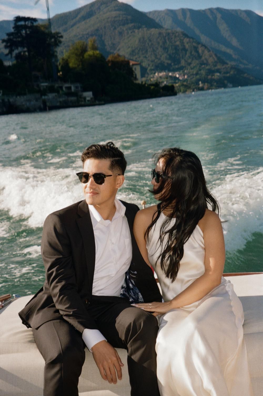 Bride and groom in sunglasses sitting on a boat with scenic mountain lake views on their wedding day.
