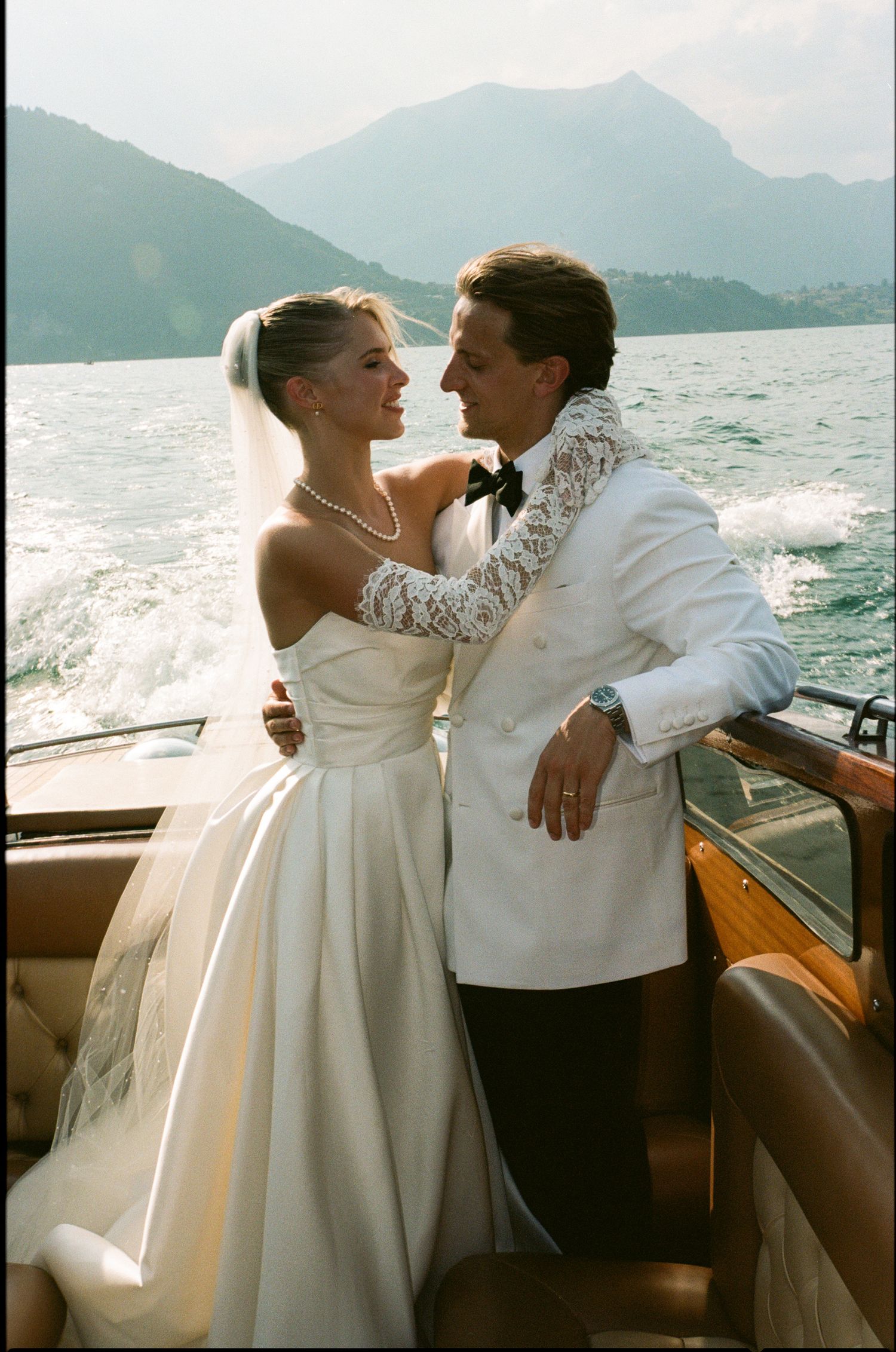 Bride in strapless gown and groom in white tuxedo embrace on classic wooden boat on mountain lake.