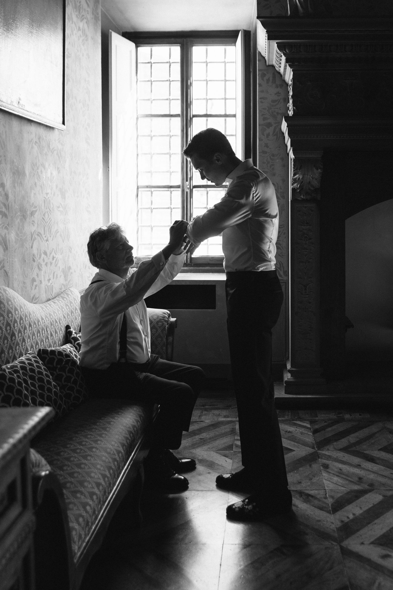 Black and white photo of two men in a historic interior, one helping the other adjust a tie by a window.