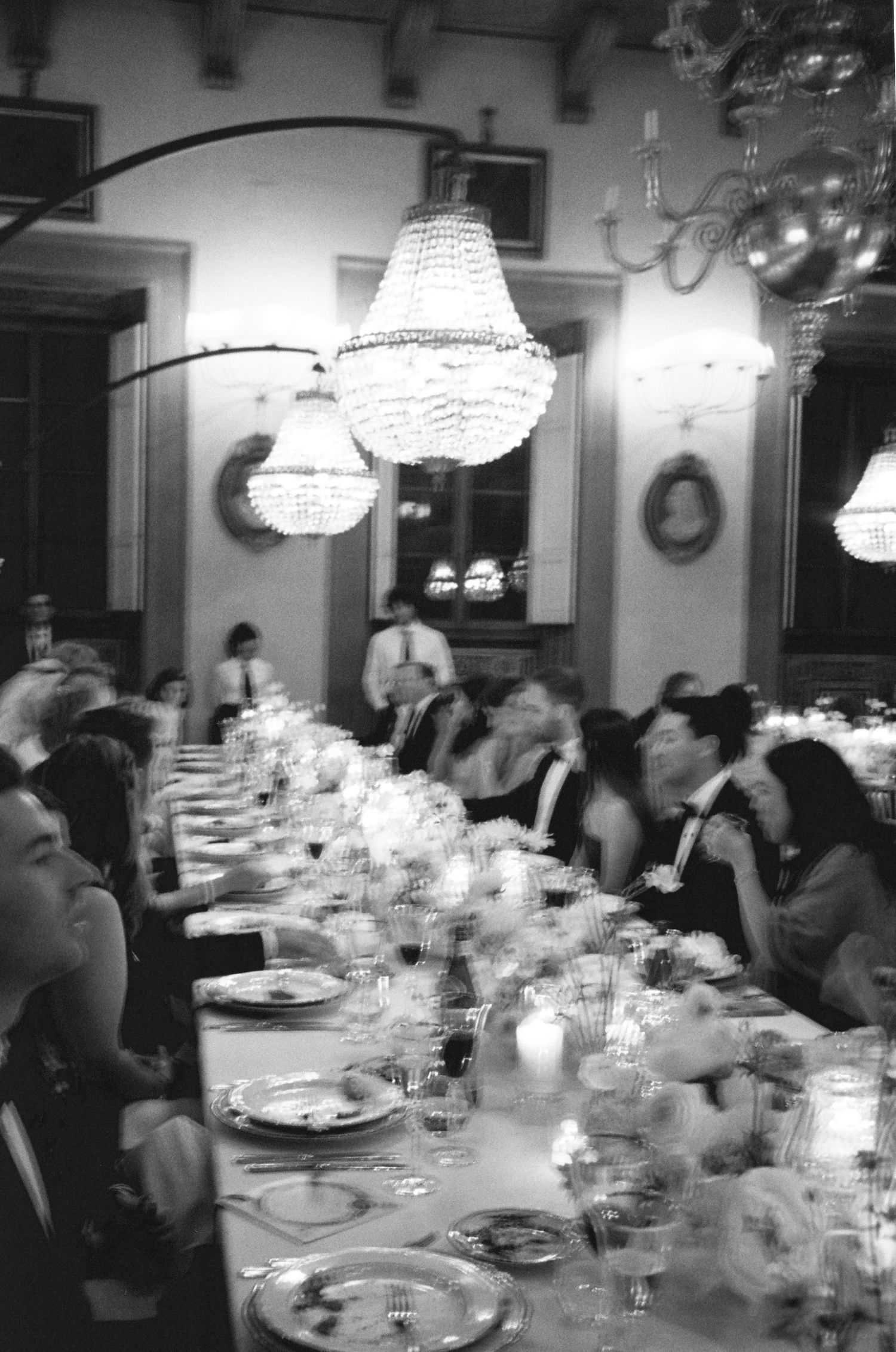 Black and white photo of guests dining under crystal chandeliers at a formal wedding reception.