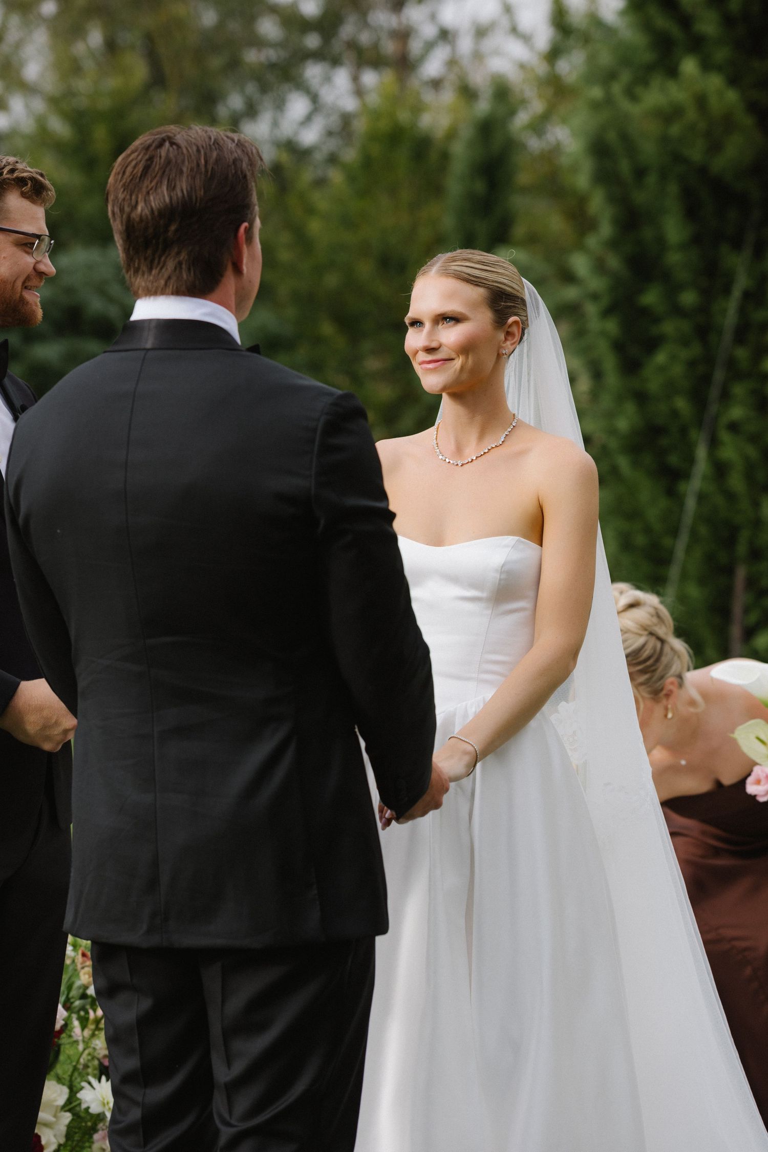 Bride in white strapless gown and veil holds hands with groom in black suit during outdoor wedding ceremony.