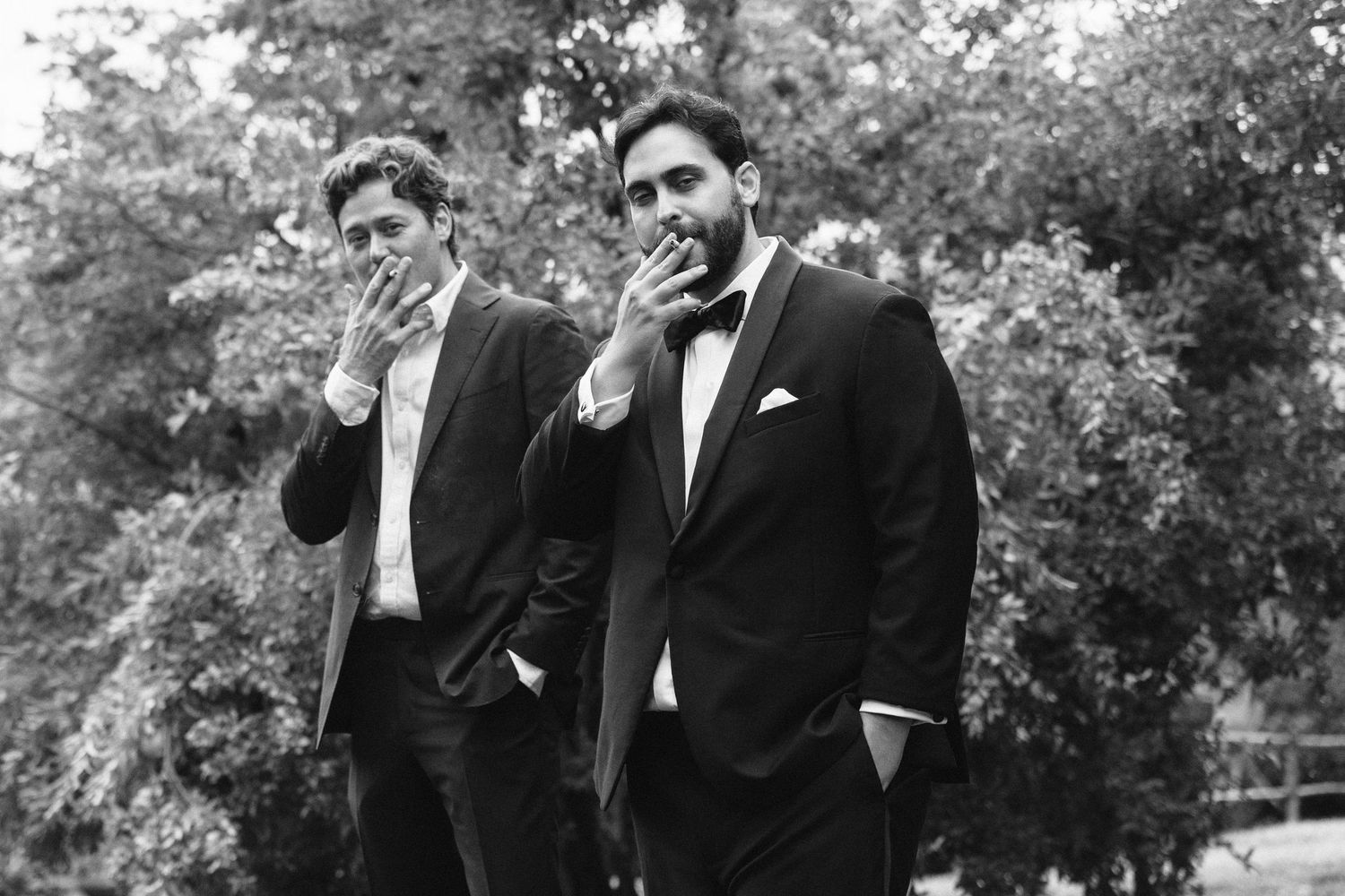 Two men in tuxedos smoking cigarettes outdoors in a black and white photo.