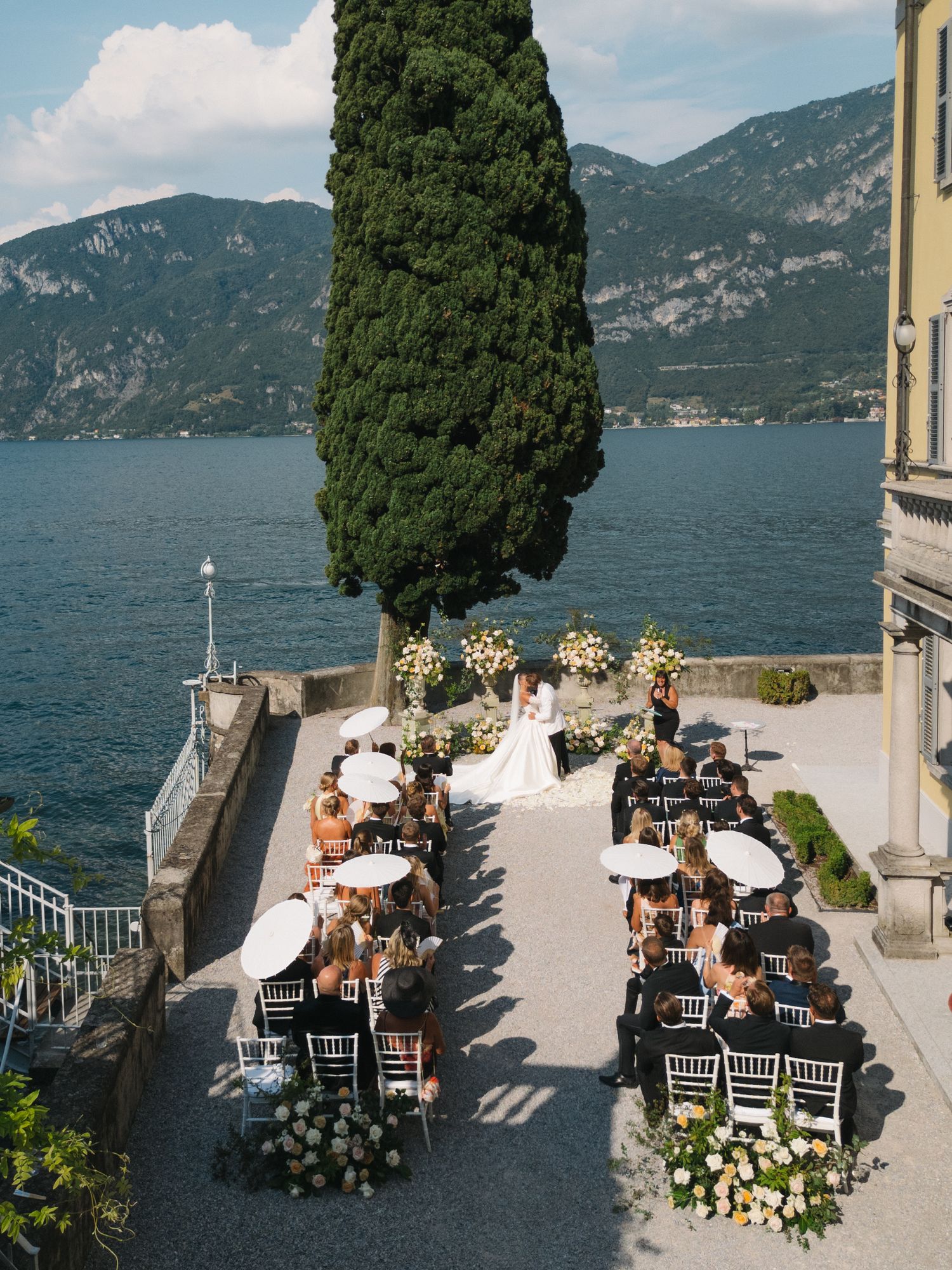Outdoor wedding ceremony on a lakeside terrace with mountain views, white parasols, and floral arrangements at Villa del Balbianello.