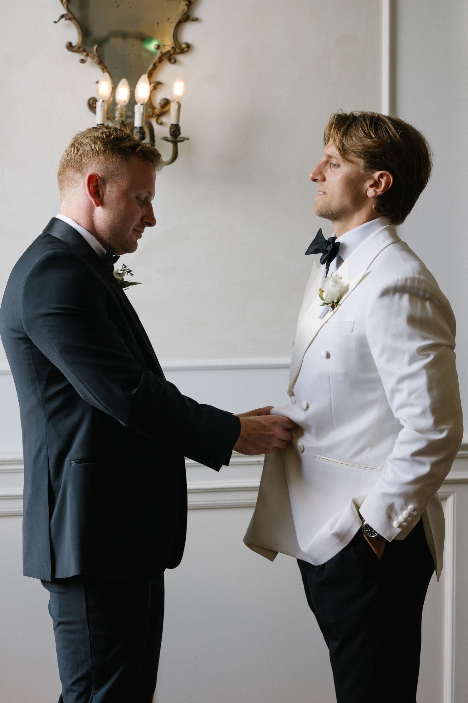 Two grooms in formal wedding attire, one in white tuxedo and one in black suit, sharing a tender moment indoors.