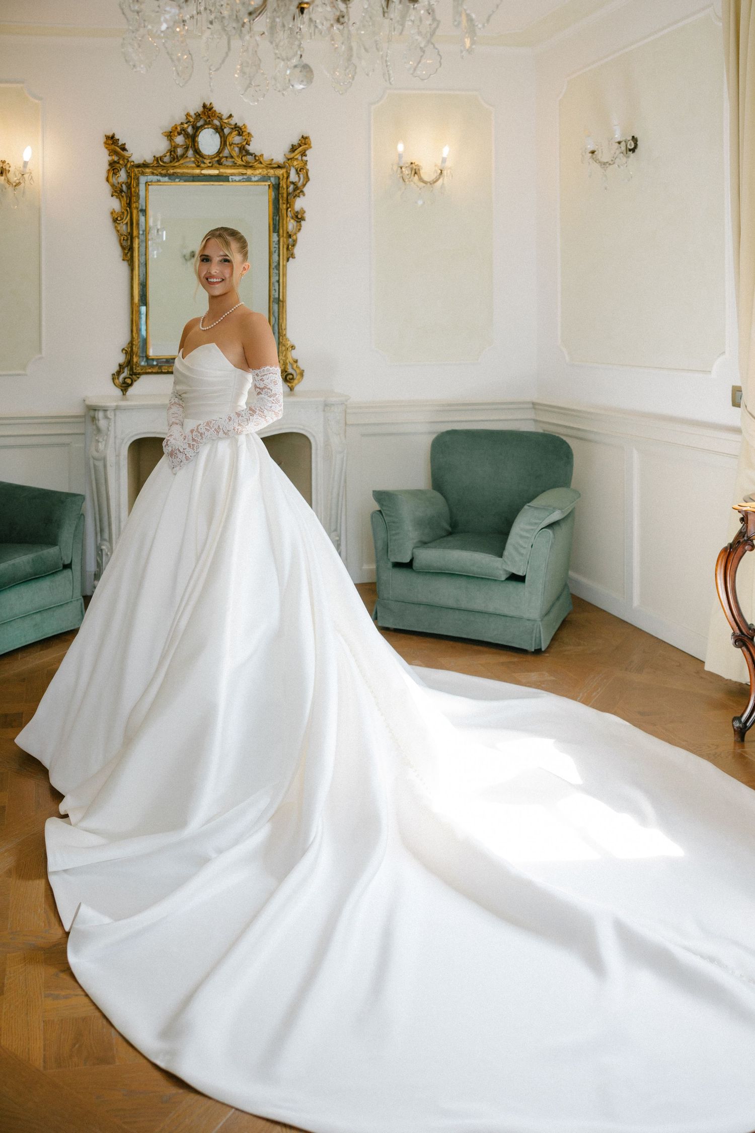 Bride in elegant white ball gown with long train poses in luxurious room with ornate gold mirror and green armchair.