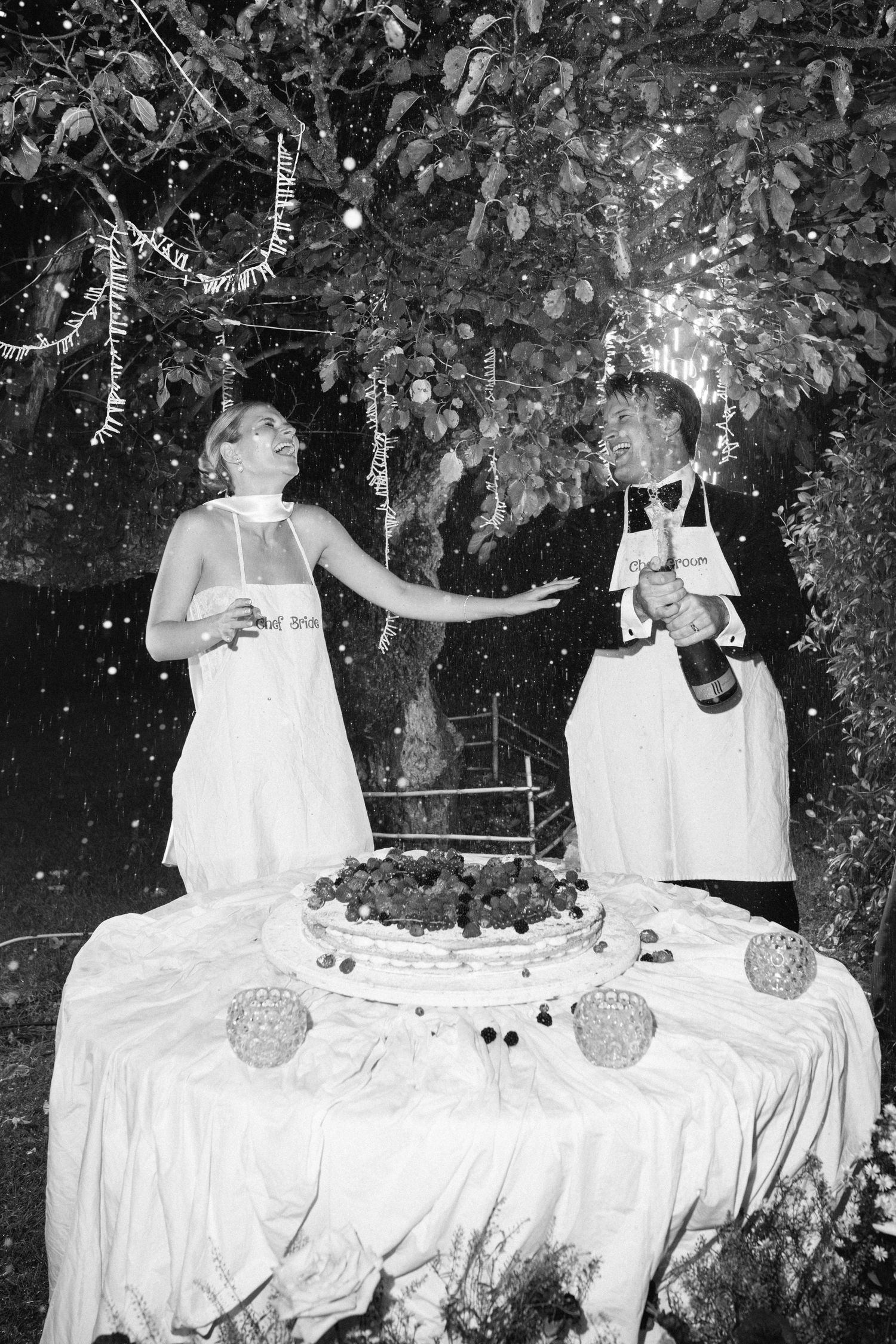 Black and white photo of a bride and groom celebrating with champagne beside a decorated wedding cake outdoors.