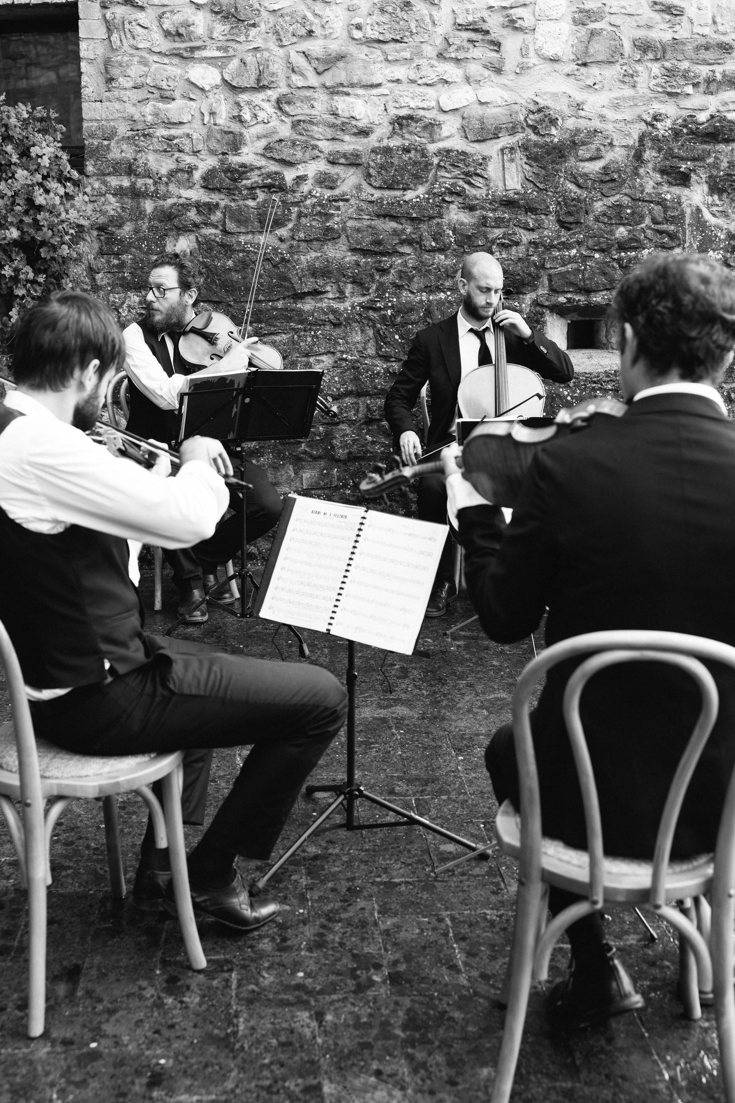 Black and white photo of a string quartet performing outdoors against a rustic stone wall with music stands.