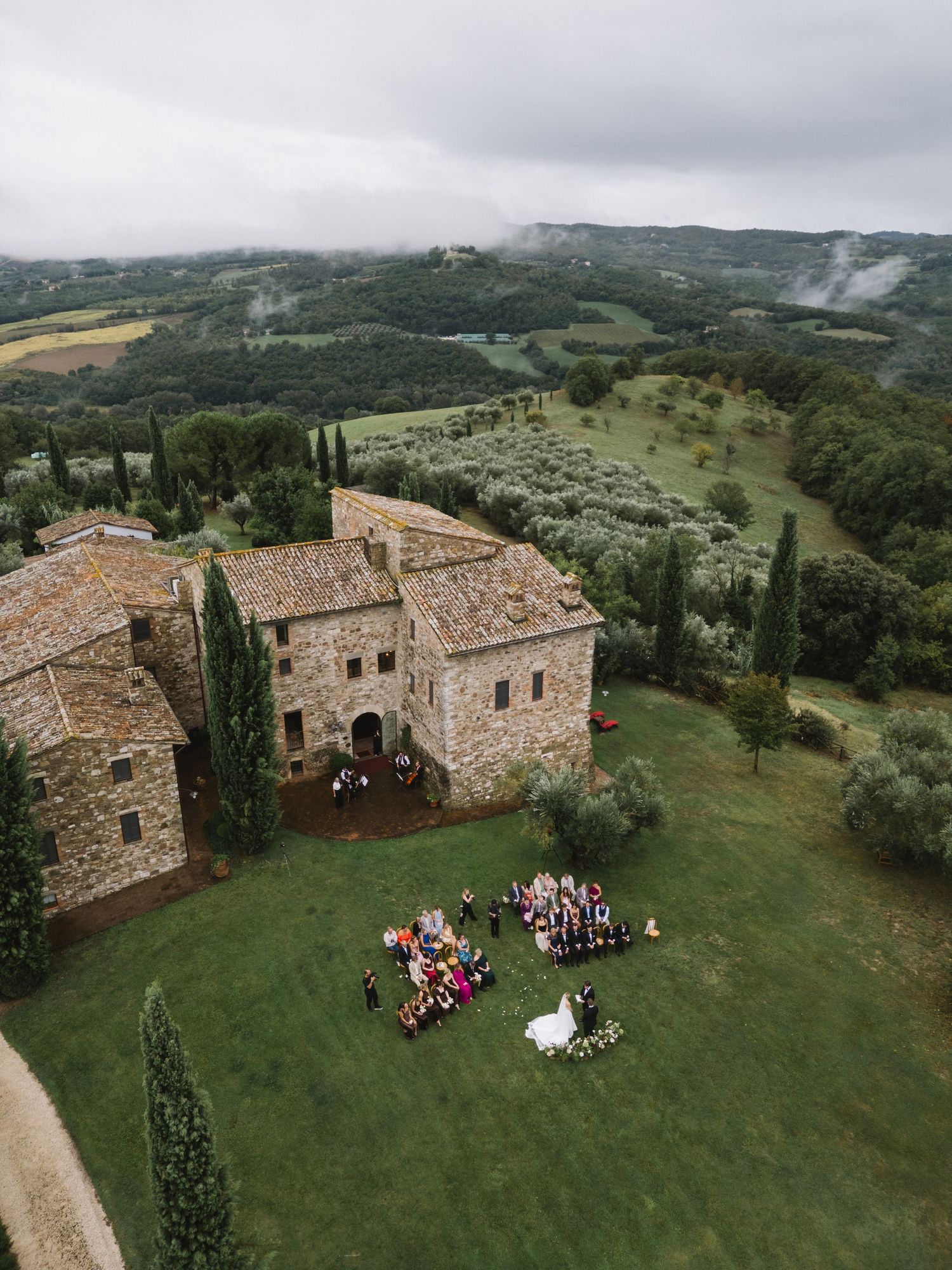 Aerial view of a Tuscan stone villa wedding ceremony surrounded by cypress trees and rolling green hills.