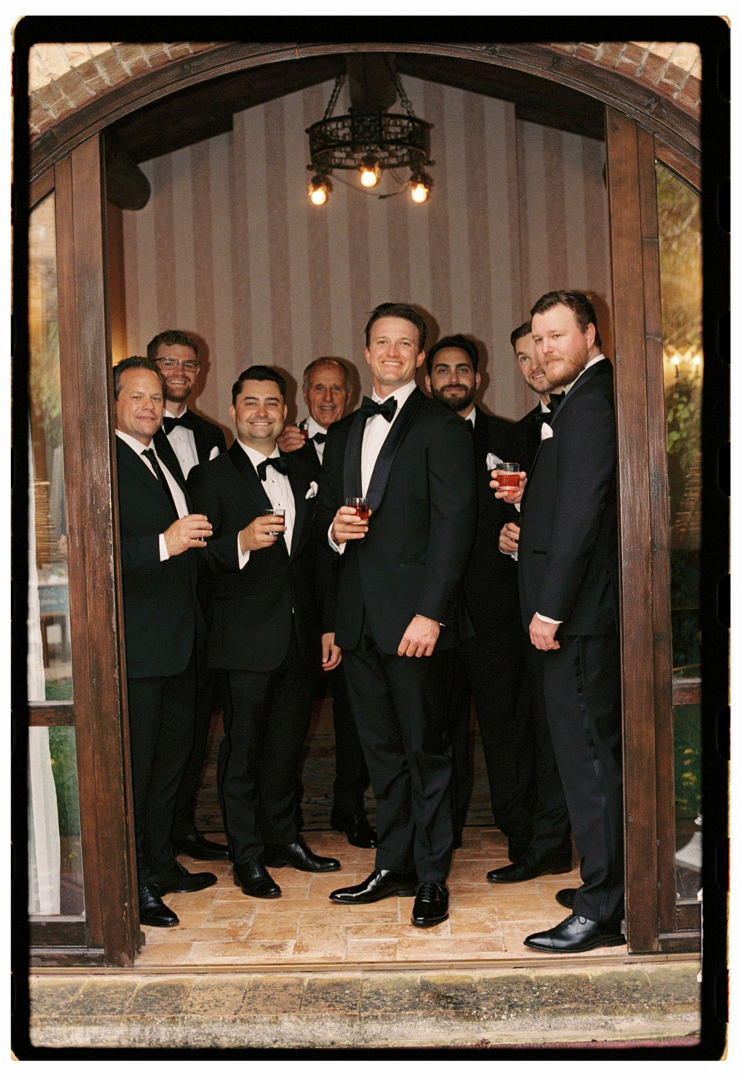 Group of groomsmen in black tuxedos holding drinks, posing together in an arched wooden doorway.