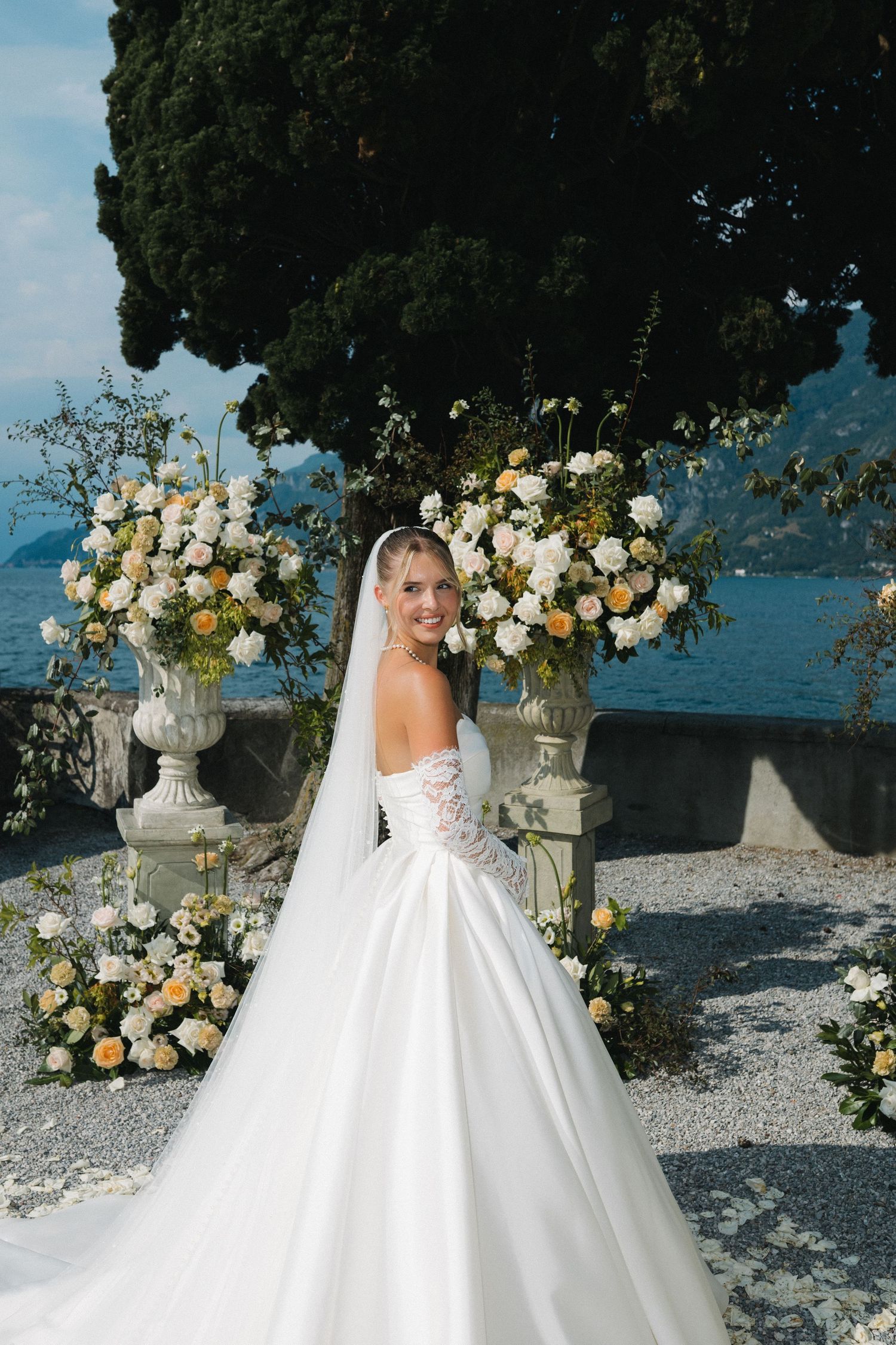 Bride in white ball gown and veil poses before floral arrangements at an elegant outdoor lakeside wedding ceremony.