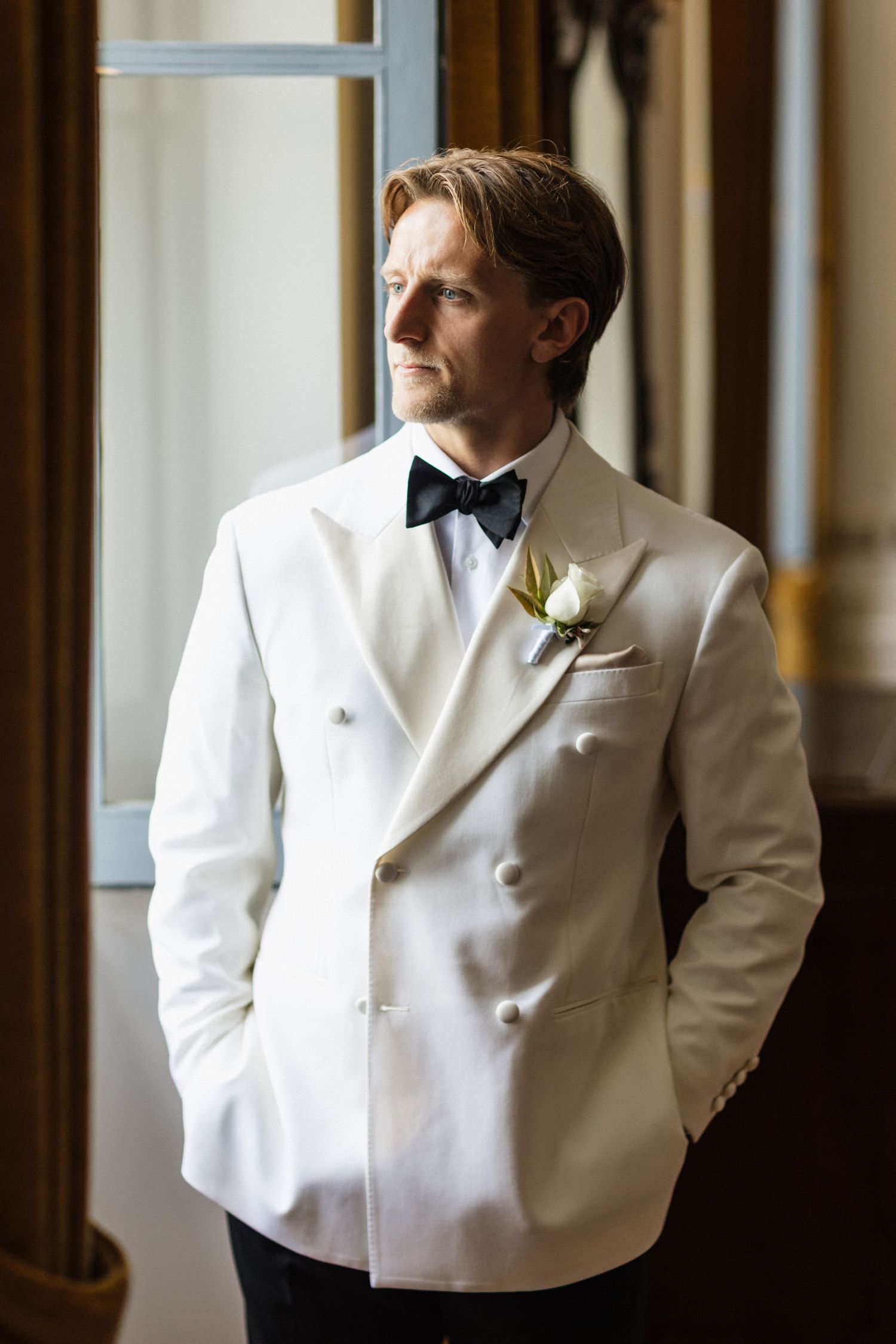 Groom in white double-breasted tuxedo jacket with black bow tie and floral boutonniere gazing out window.