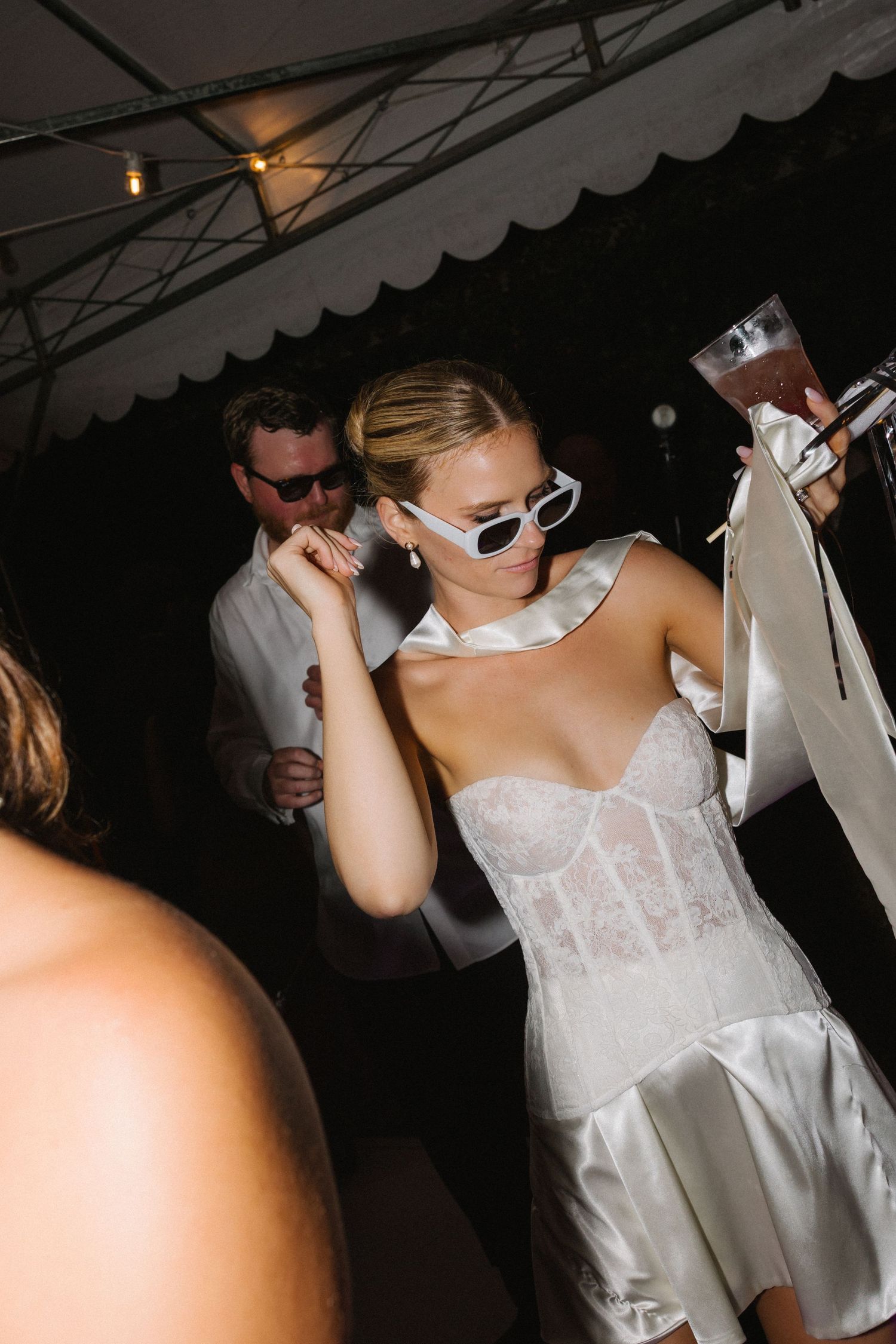 Bride in white lace corset dress and white sunglasses dancing at wedding reception under tent.