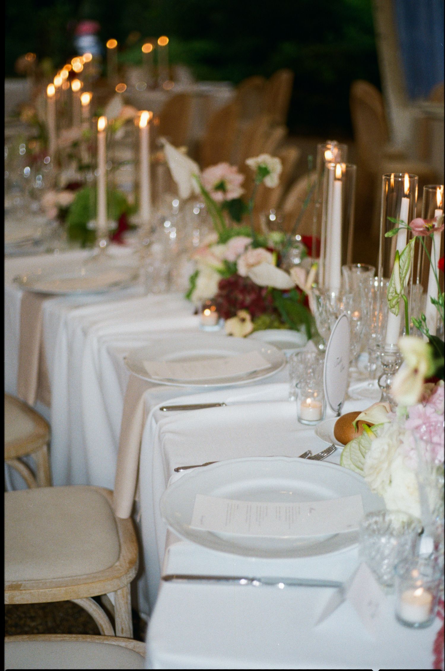 Elegant wedding reception table with white linens, floral centerpieces, candles, and place settings in a romantic evening setting.