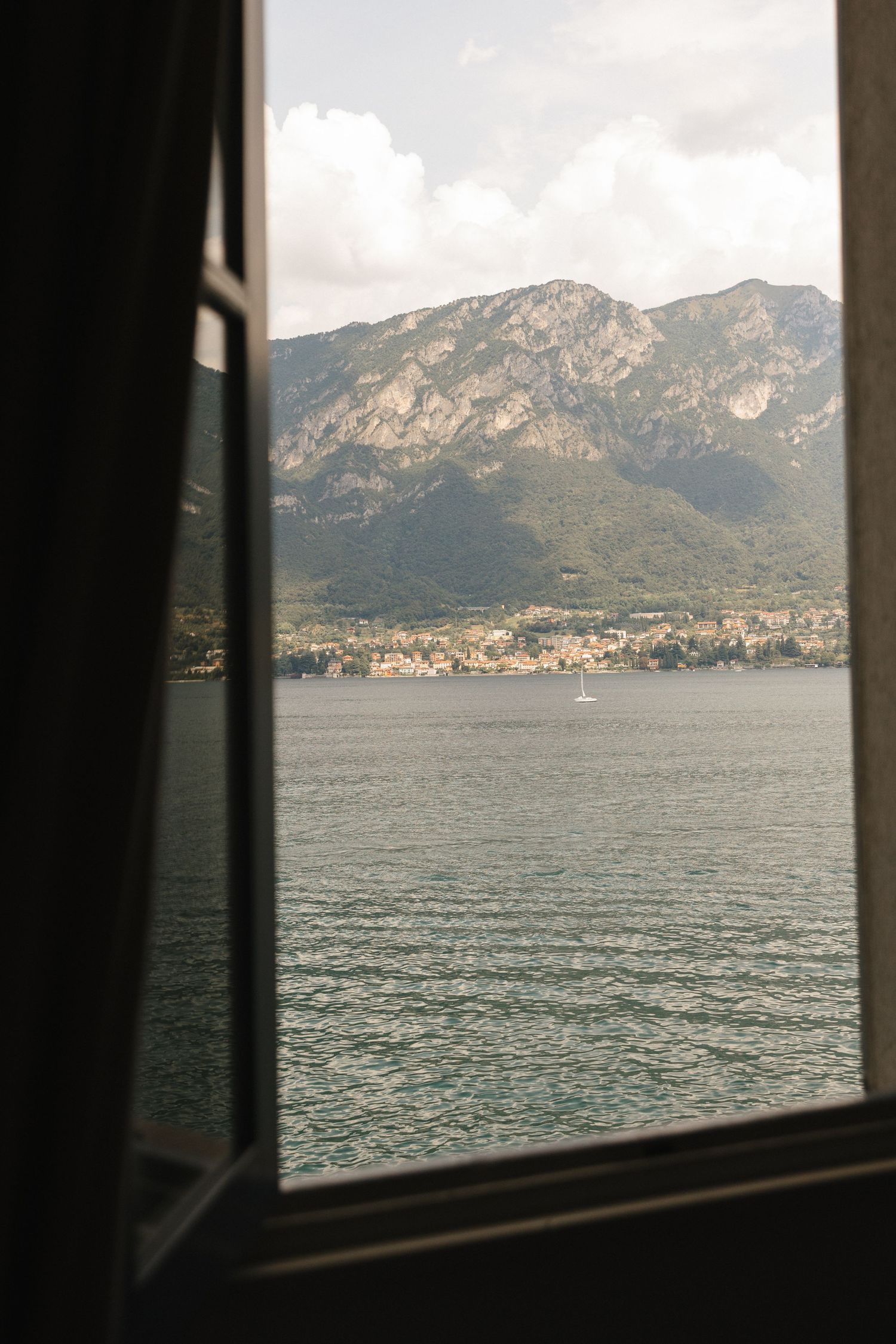 View through open window of serene lake with mountain backdrop and small town on shore under cloudy sky.