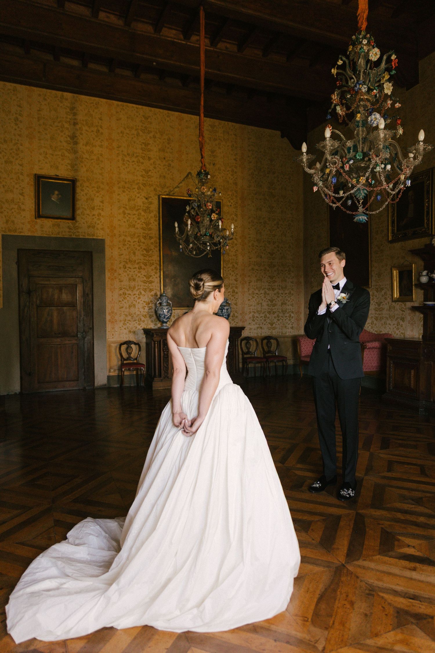 Bride in strapless ball gown with train stands before emotional groom in chandelier-lit ornate ballroom.