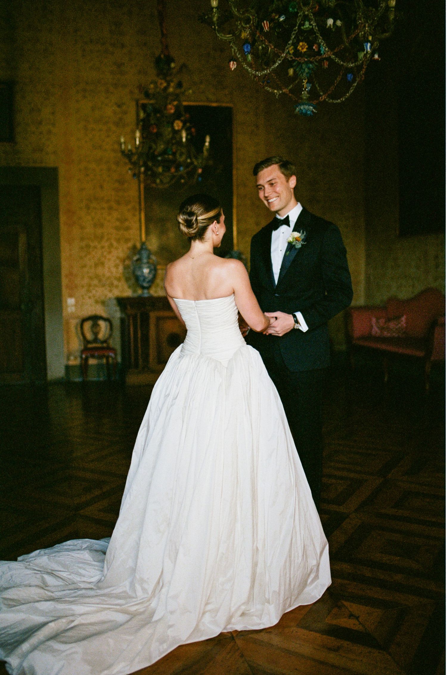Bride in strapless ball gown and groom in black tuxedo share romantic moment in ornate historic room.