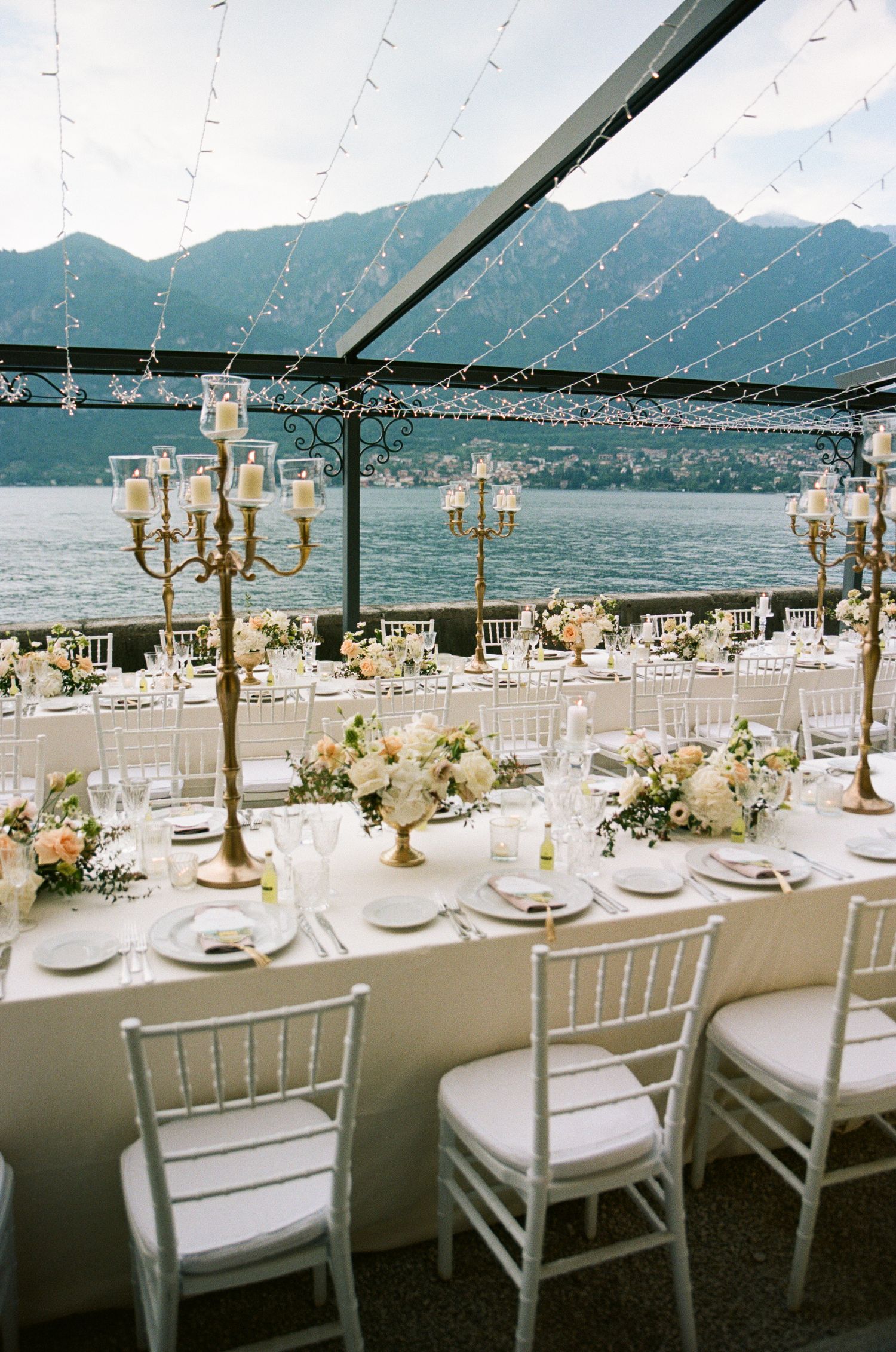 Elegant lakeside wedding reception with gold candelabras, white florals, and string lights over water.