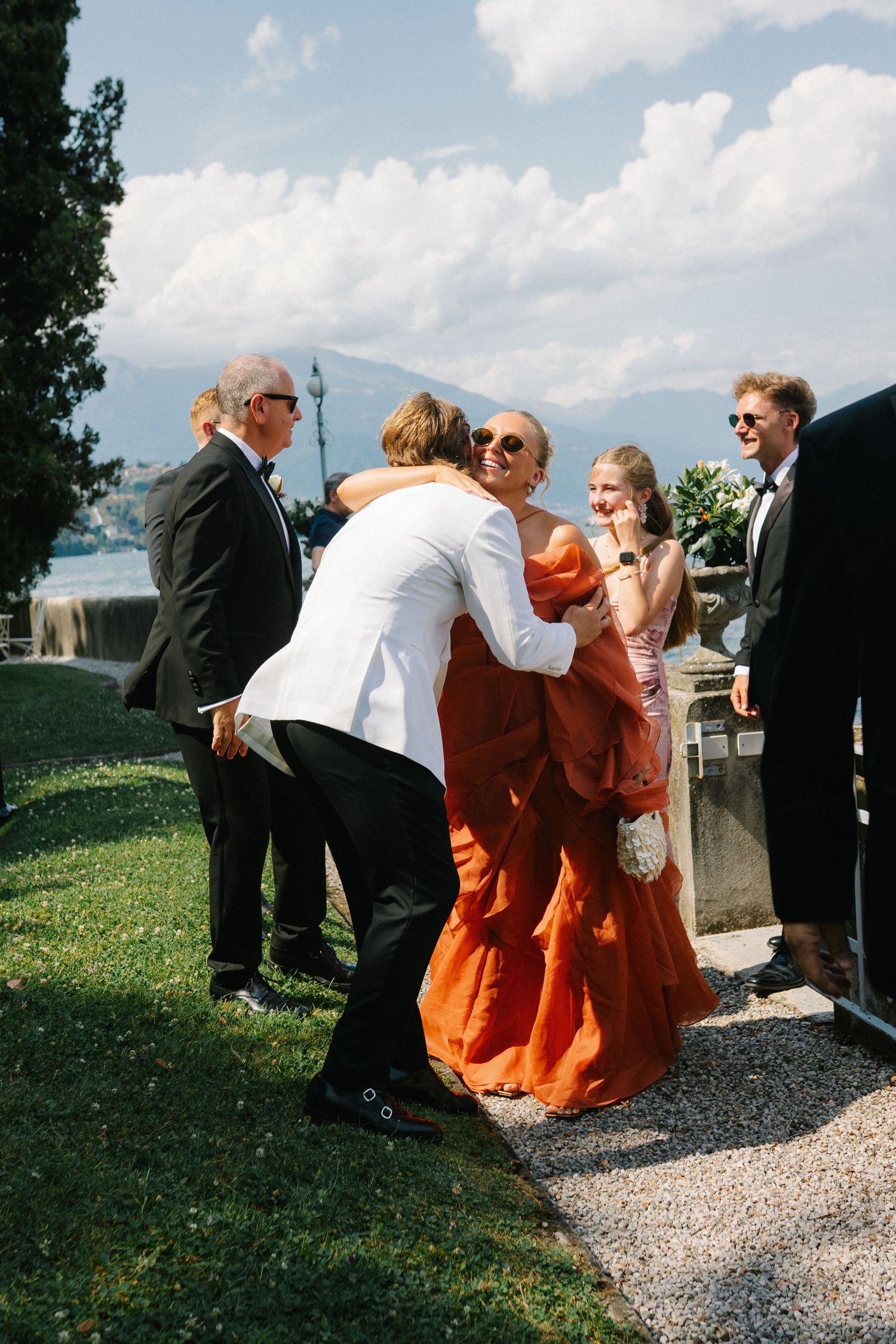 Guests in formal attire embrace at an outdoor wedding reception with mountain lake views on a sunny day.