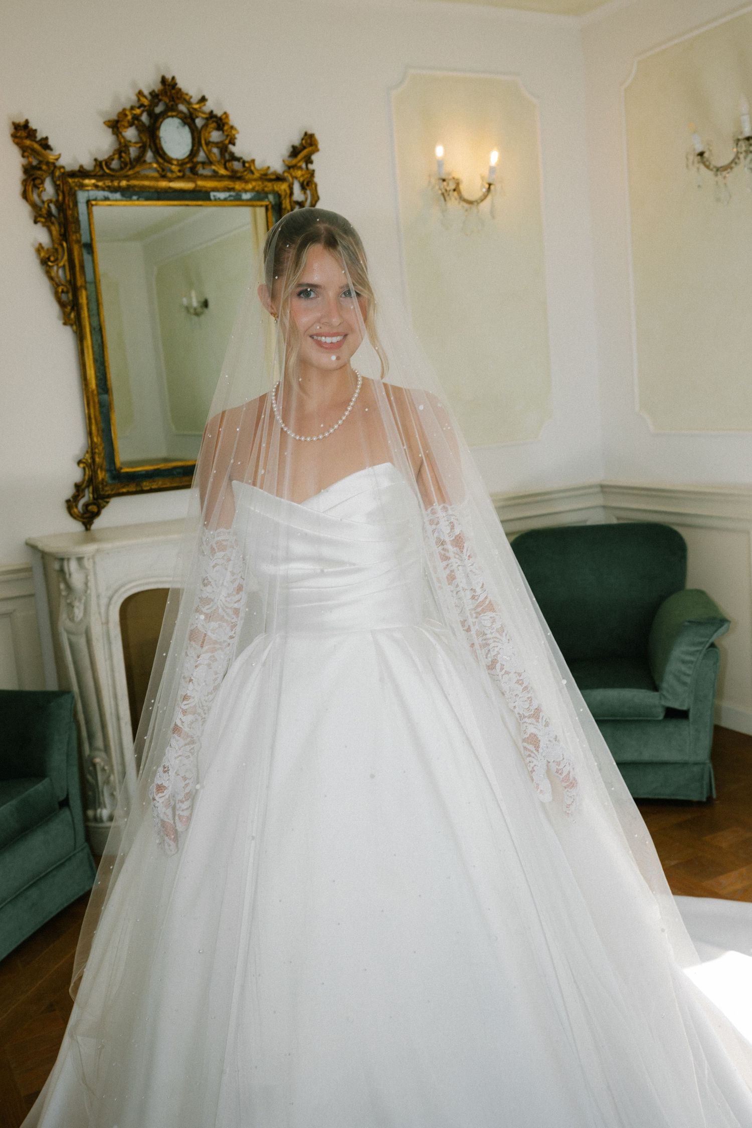 Smiling bride in a white ball gown and lace veil stands in an elegant room with a gold mirror and green velvet chairs.