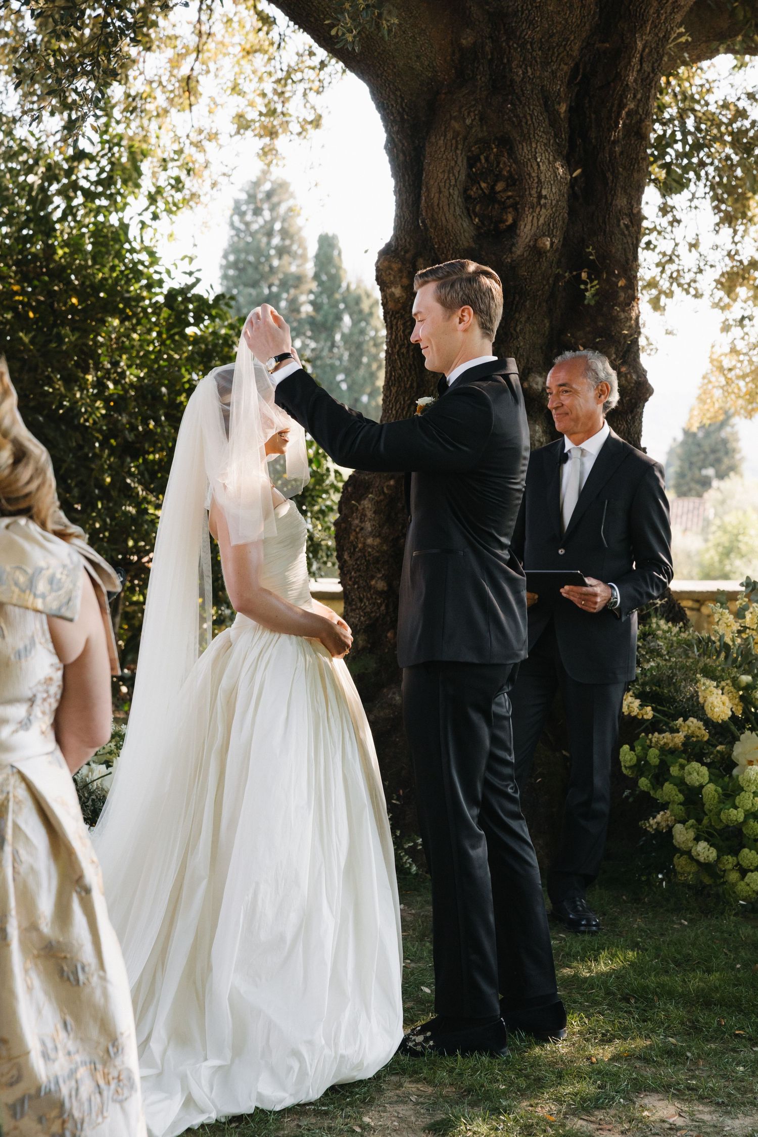 Groom lifting bride's veil during outdoor wedding ceremony beneath ancient olive tree.