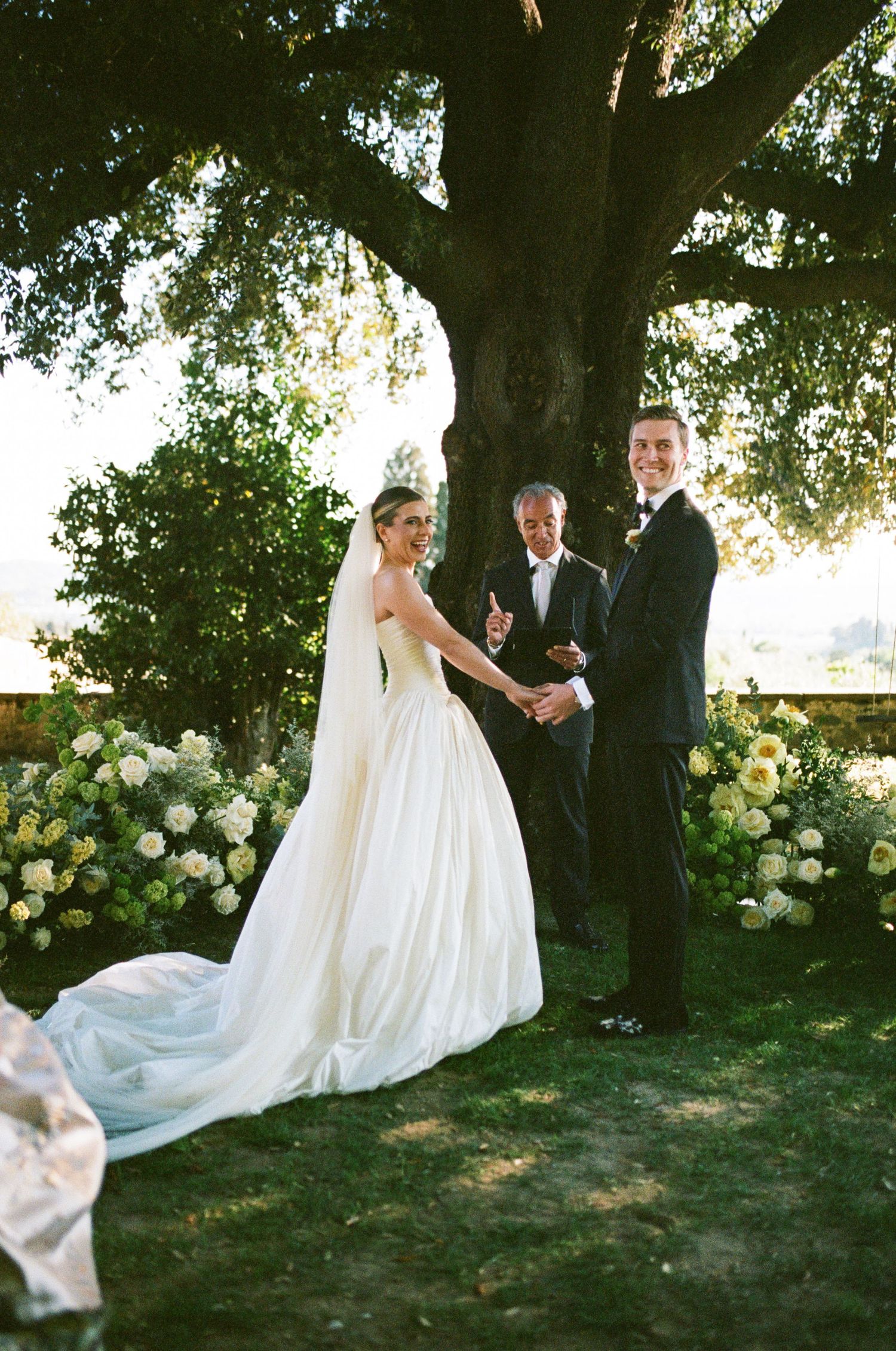 A bride and groom exchange vows outdoors beneath a large tree surrounded by white and yellow floral arrangements.