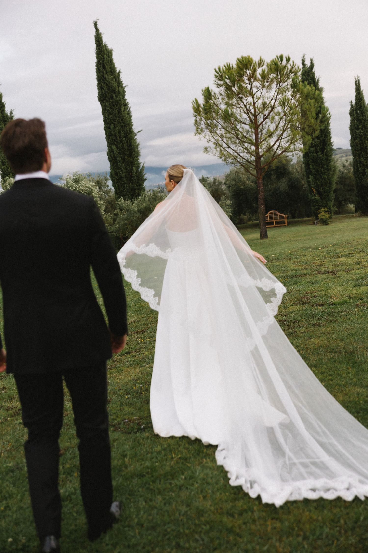 Bride in flowing white gown and veil walks with groom in black suit through lush green garden with cypress trees.