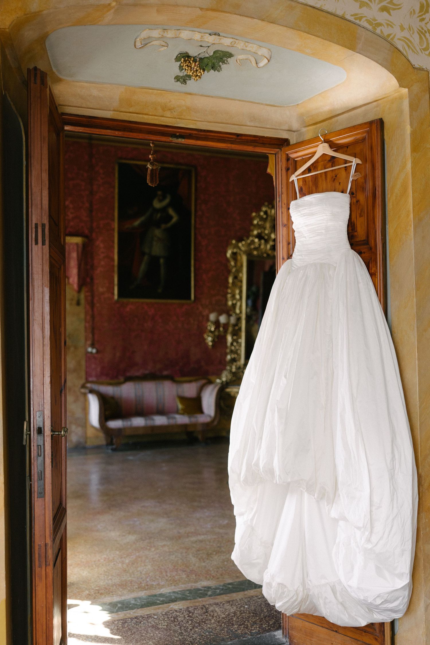 White wedding dress hanging on wooden hanger in doorway of elegant vintage room with ornate decor.