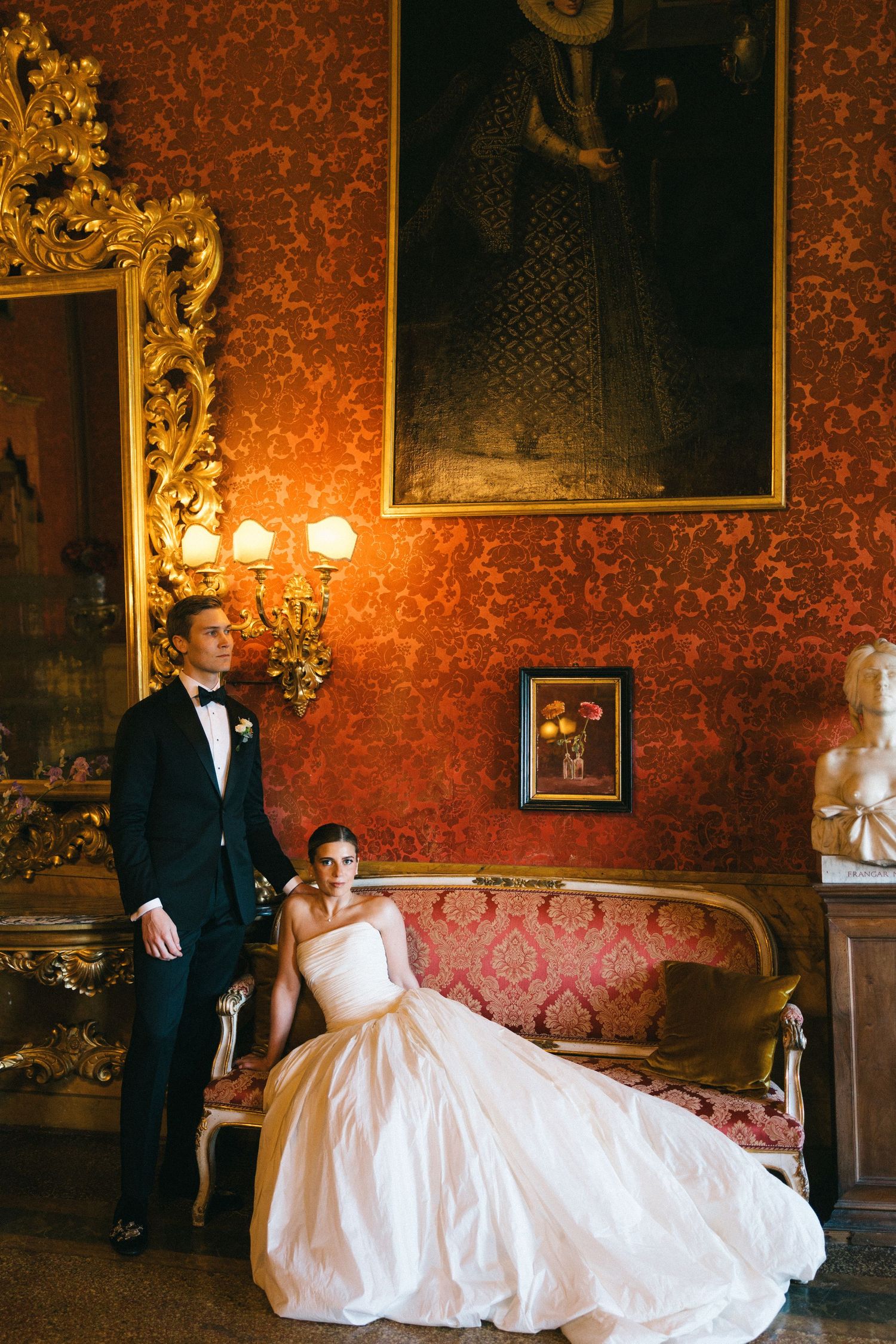 Elegant bride in white gown sits on ornate sofa beside groom in tuxedo in a luxurious red damask room.