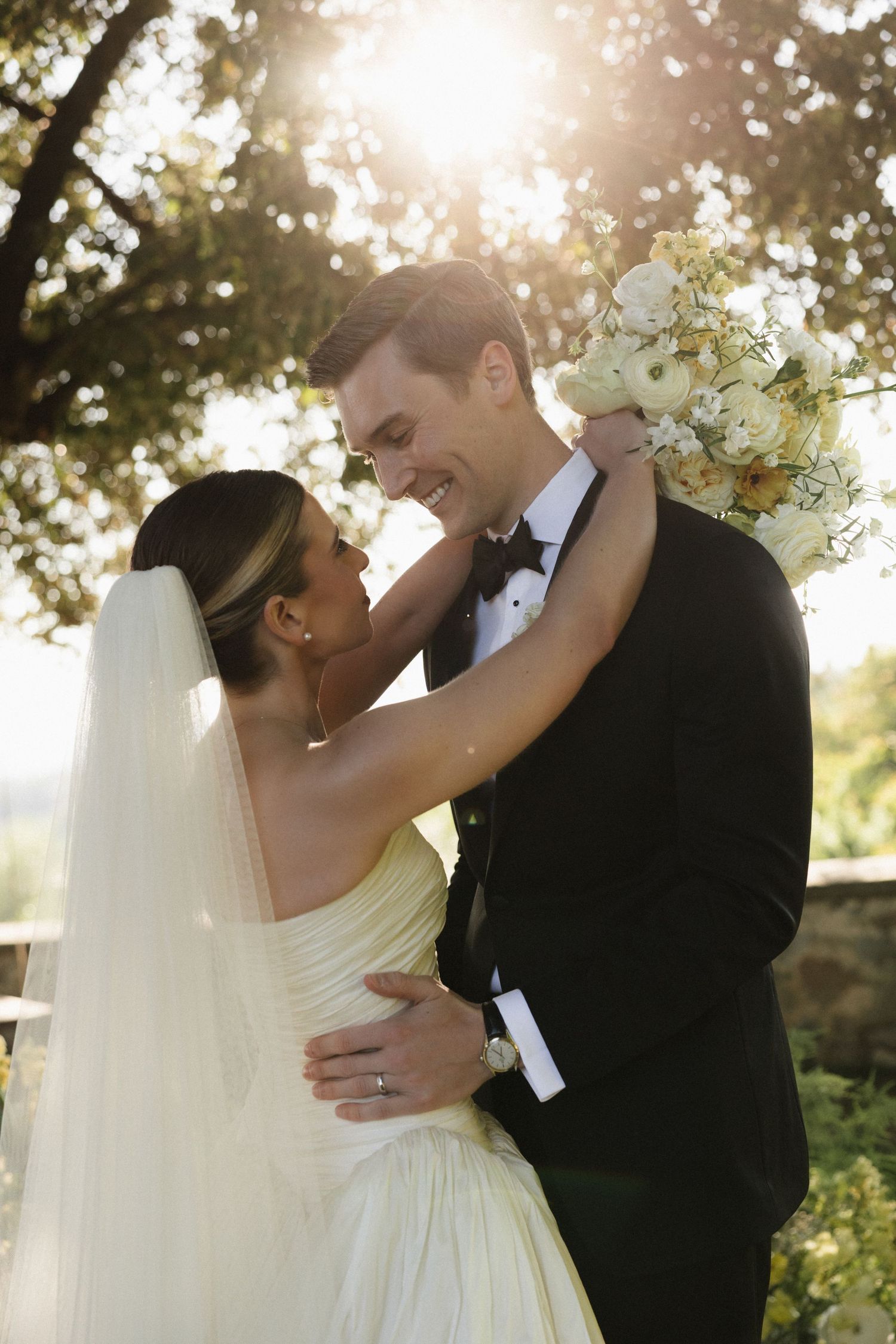Bride and groom embrace outdoors in golden sunlight, she in a white gown with veil, he in a black tuxedo holding a white bouquet.