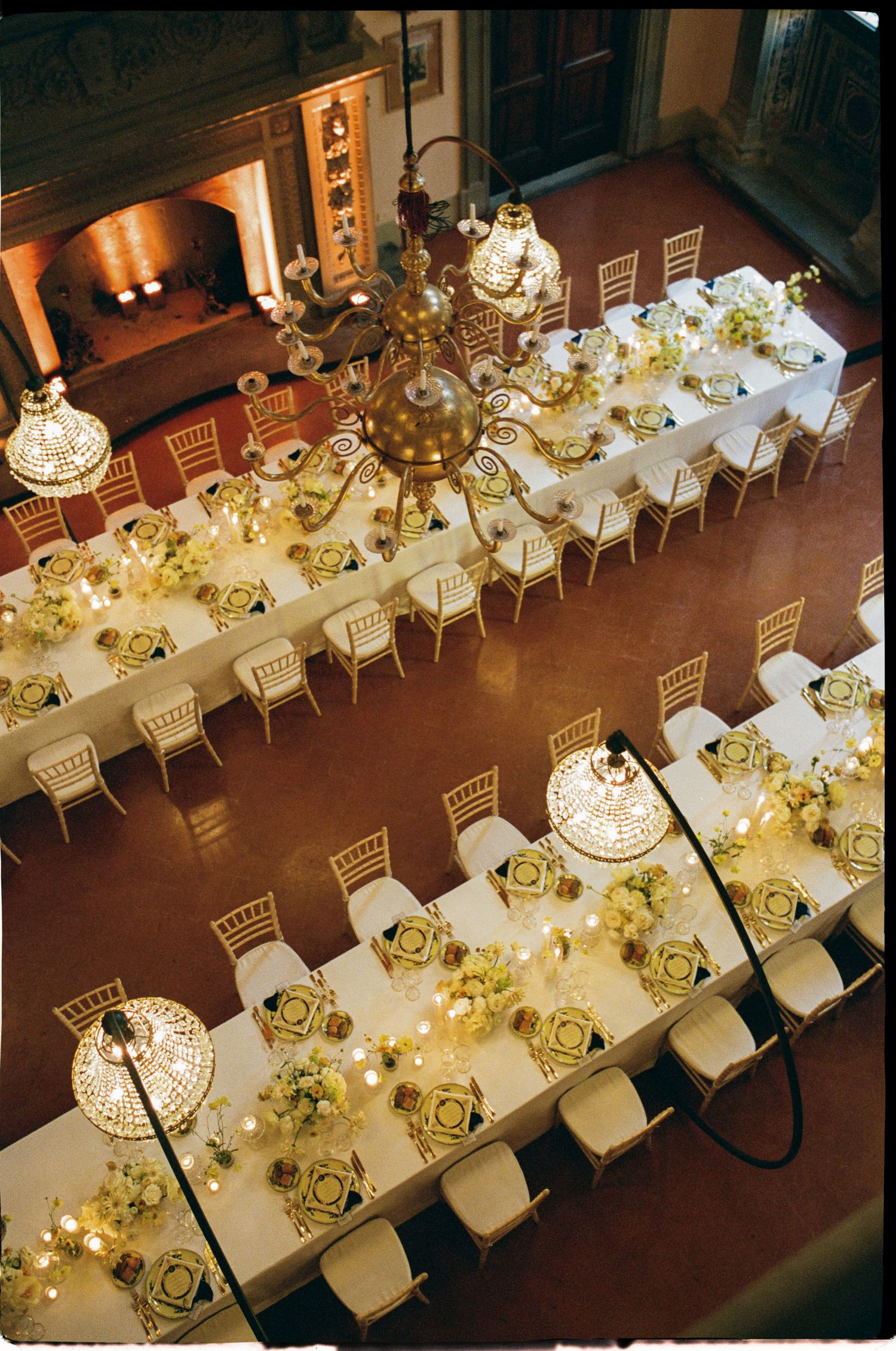 Aerial view of elegant wedding reception with long white tables, gold chandeliers, and floral centerpieces in a grand hall.