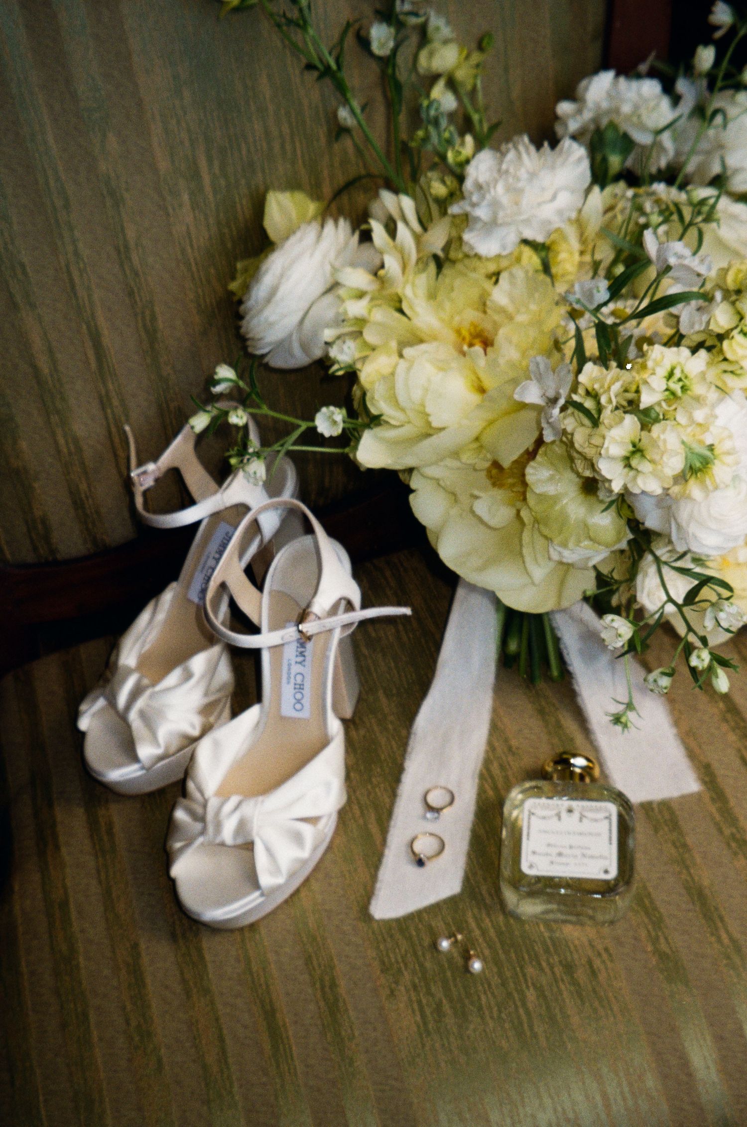 White Jimmy Choo wedding heels beside a yellow and white bridal bouquet, gold rings, pearl earrings, and perfume bottle.