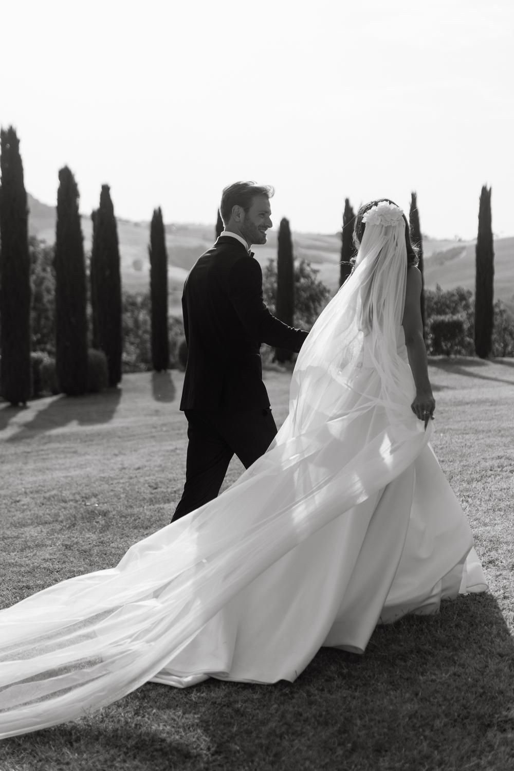 Bride and groom walking hand in hand through a Tuscan landscape lined with cypress trees in a black and white photo.