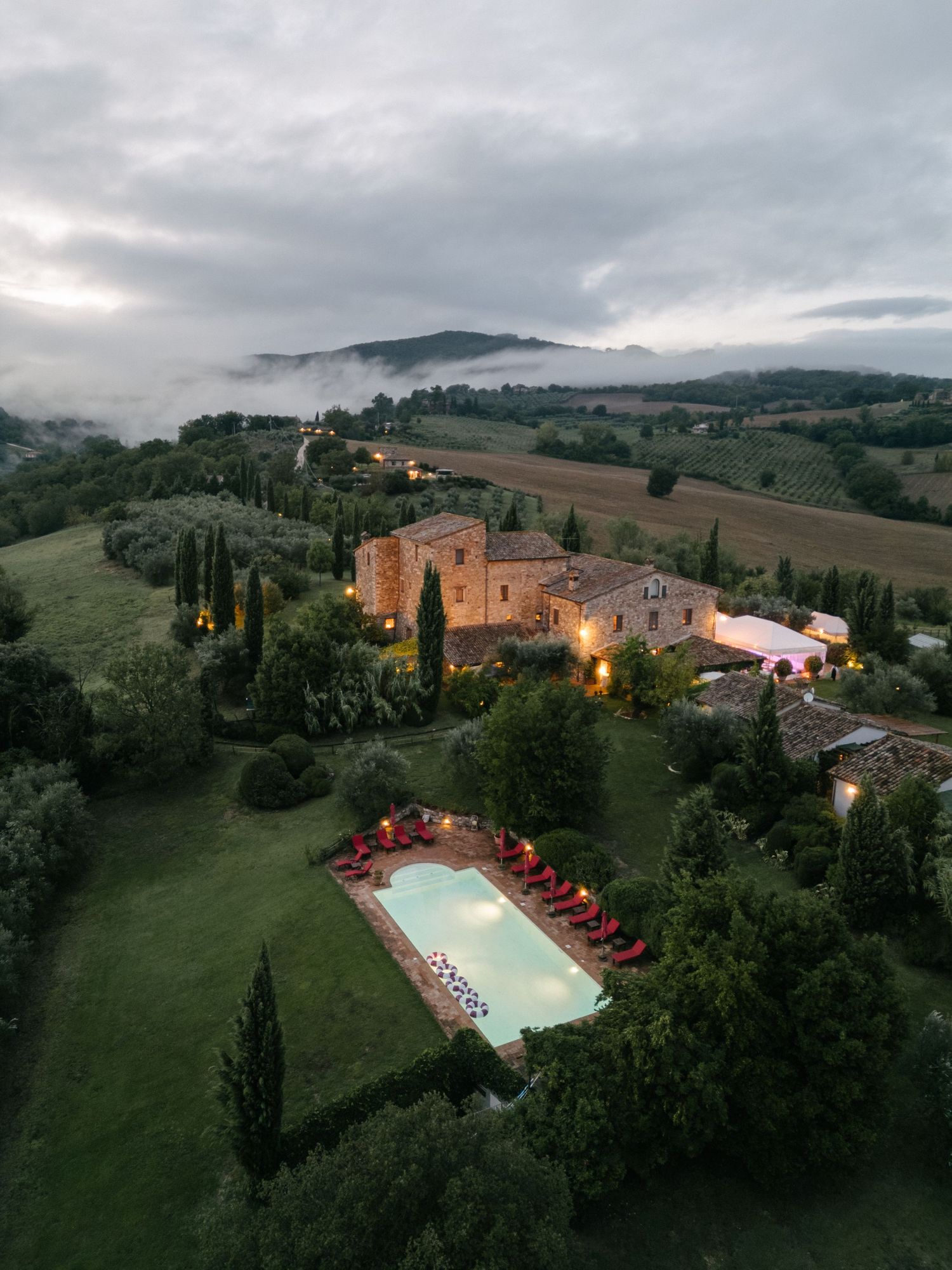 Aerial view of a Tuscan stone villa with illuminated pool, cypress trees, and misty rolling hills at dusk.