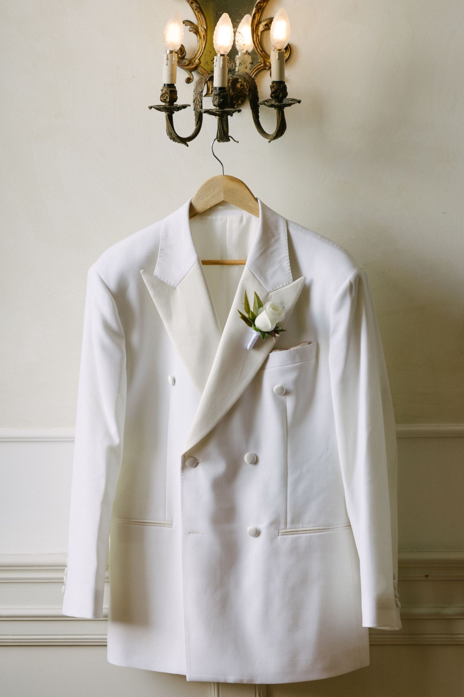White double-breasted wedding tuxedo jacket with boutonniere hanging on wooden hanger beneath vintage gold candelabra wall sconce.