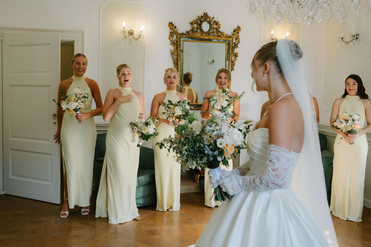 Bride in white gown reveals dress to bridesmaids in ivory dresses holding bouquets in elegant gold-accented room.