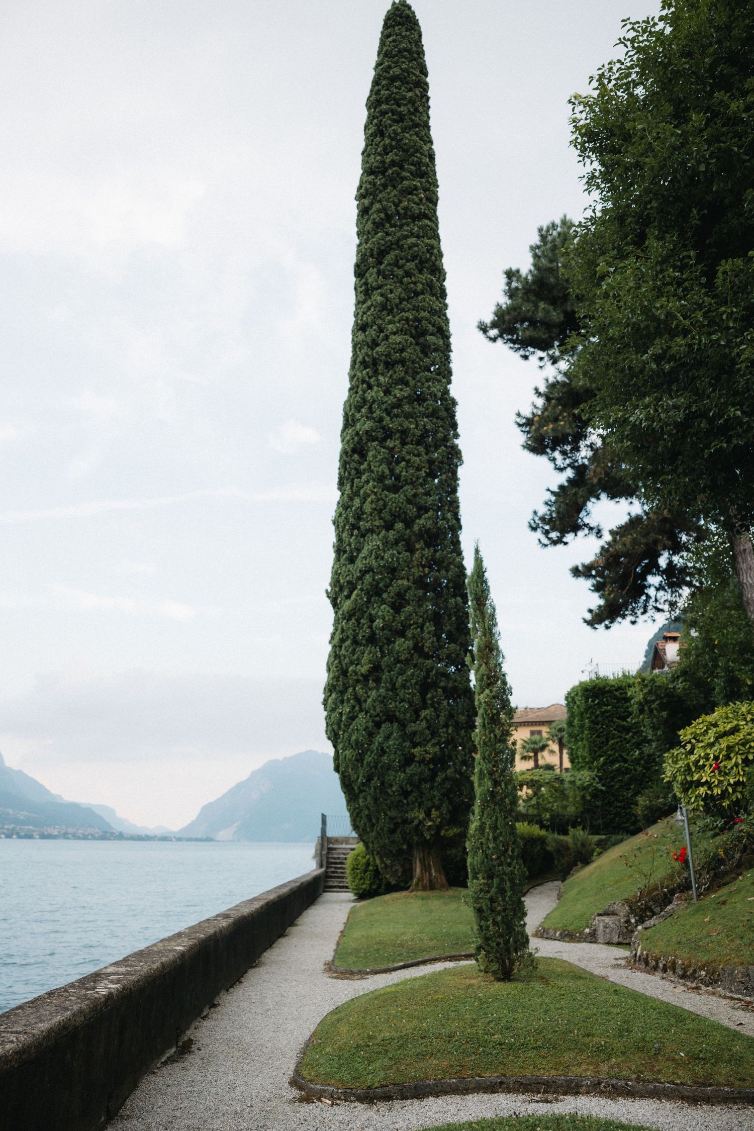 Tall cypress trees line a lakeside garden path with mountain views and a yellow villa in the background.