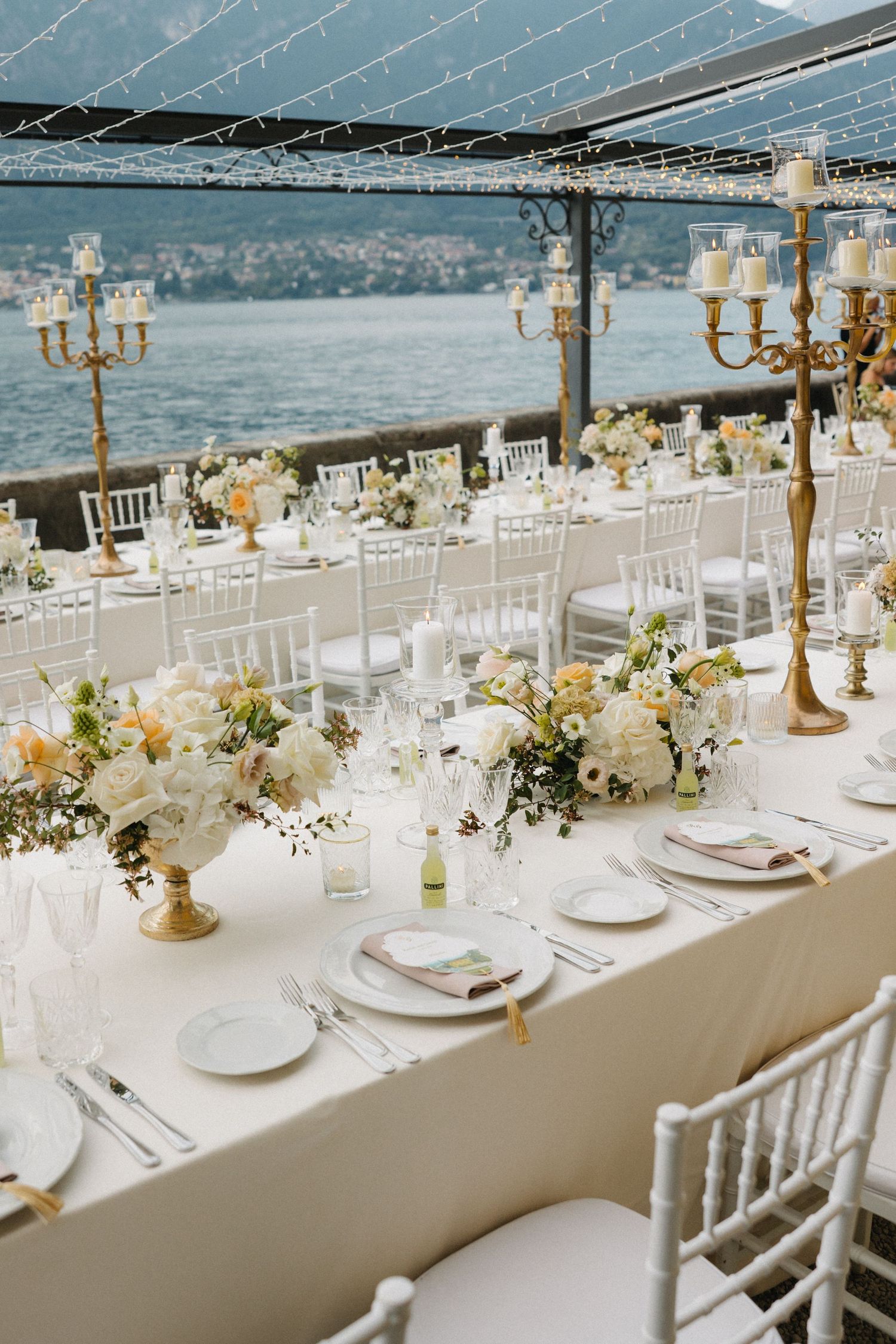 Elegant waterfront wedding reception with white Chiavari chairs, gold candelabras, and white floral centerpieces on long draped tables.