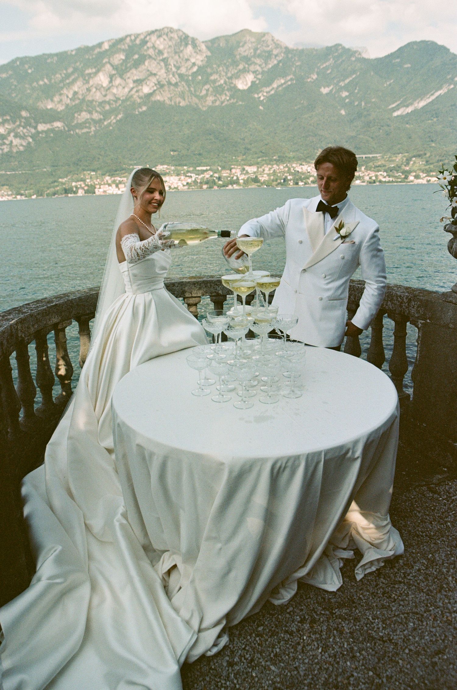 Bride and groom pouring champagne tower on scenic Lake Como balcony with mountain backdrop at luxury Italian wedding.