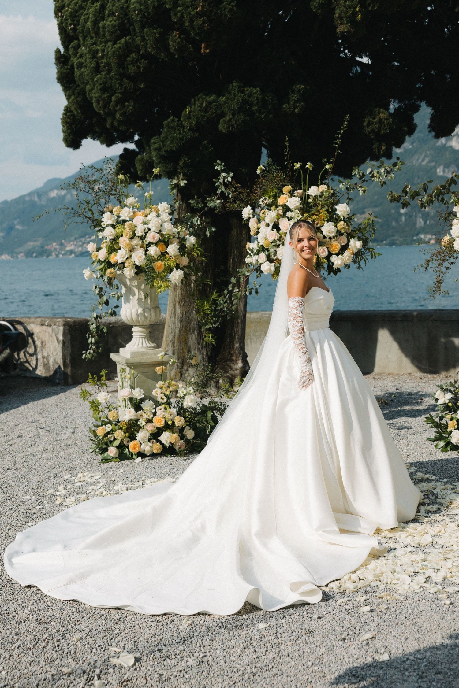 Bride in elegant white ball gown with lace gloves poses by floral arrangements overlooking a serene lake.