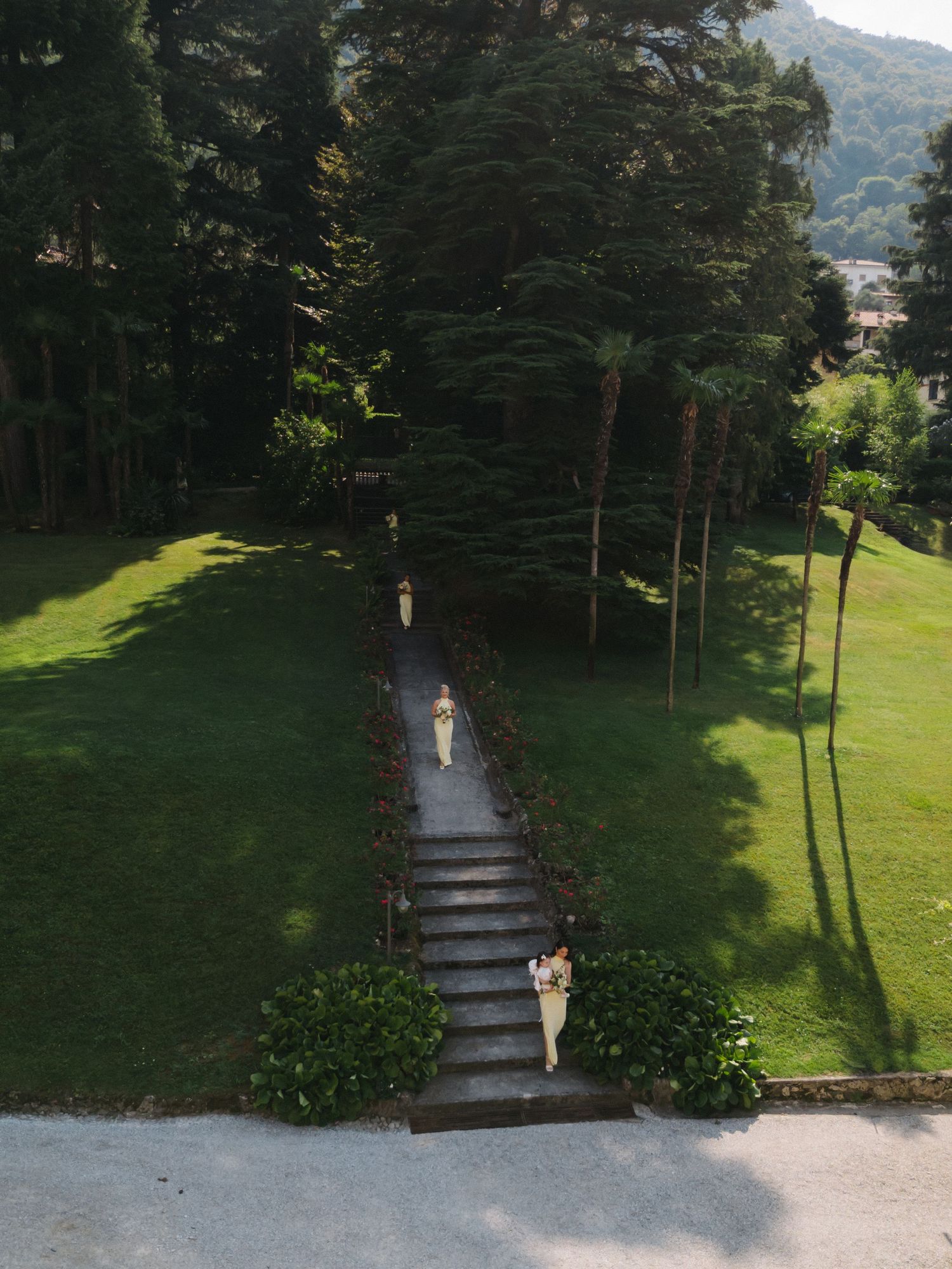 Aerial view of a lush garden path with stone steps and tall trees, two people walking through manicured grounds.
