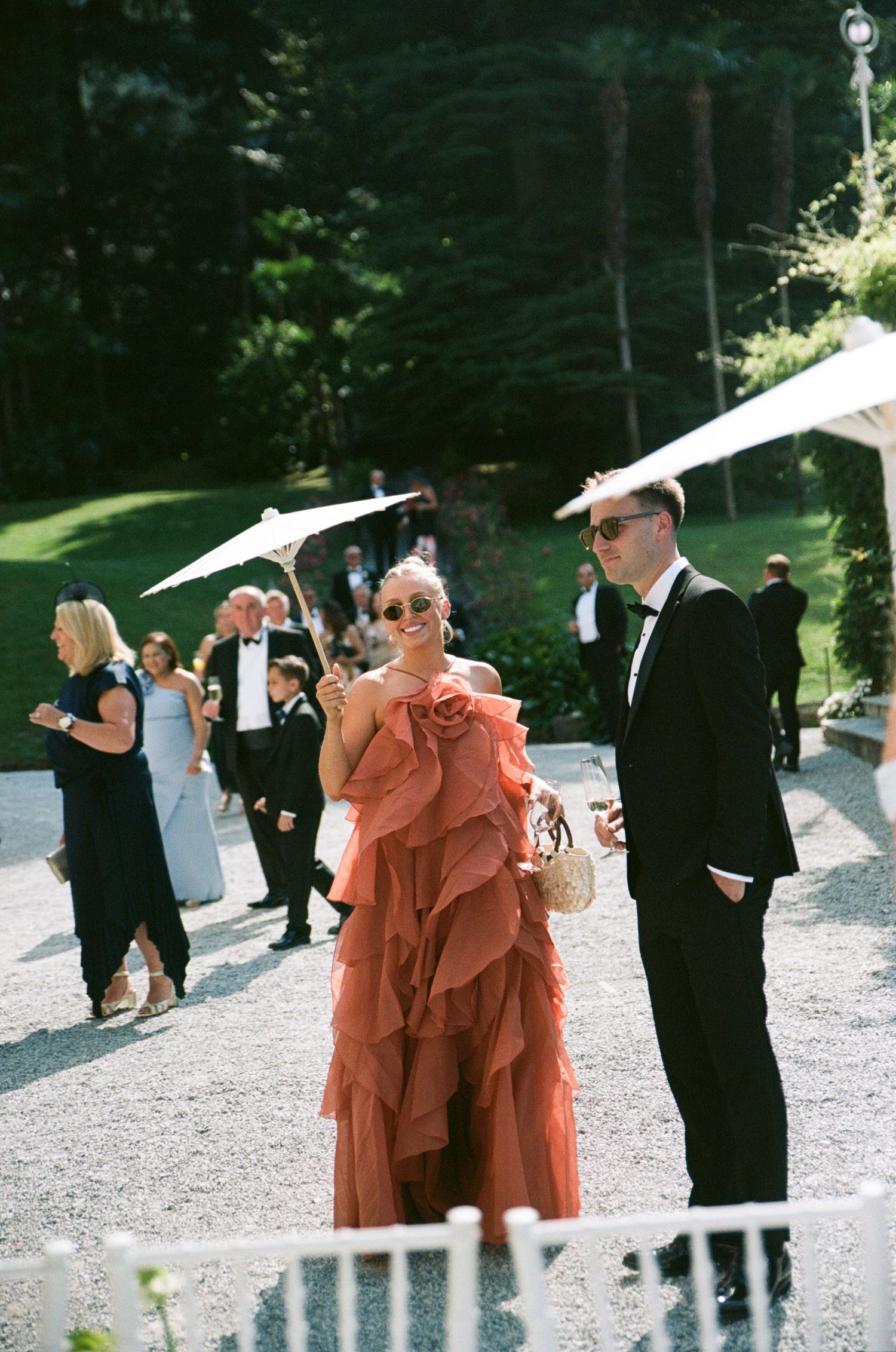 Woman in ruffled coral gown and man in tuxedo walk at elegant outdoor wedding reception with guests in background.