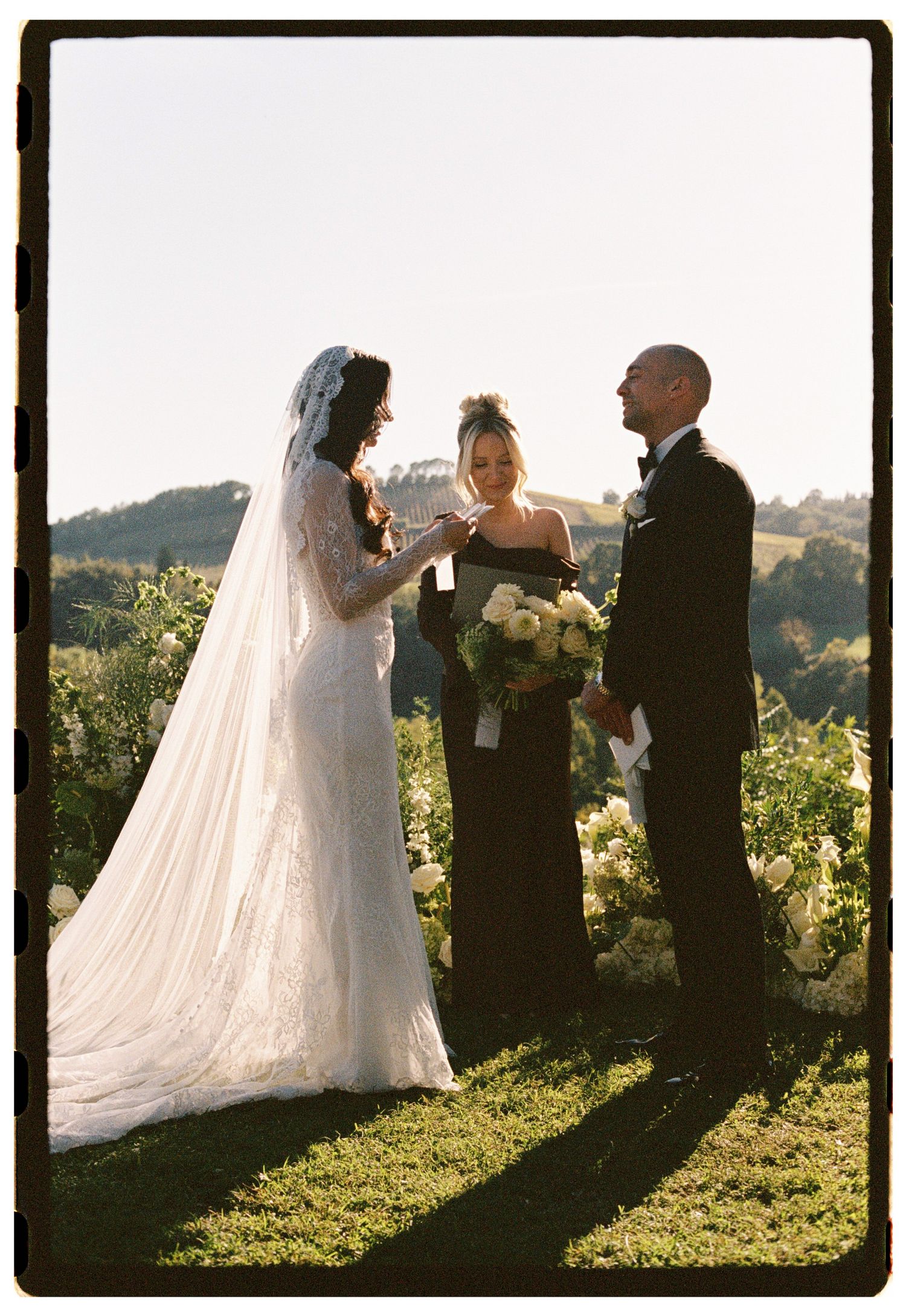 Bride and groom exchange vows outdoors with officiant, surrounded by white florals and rolling hills at golden hour.