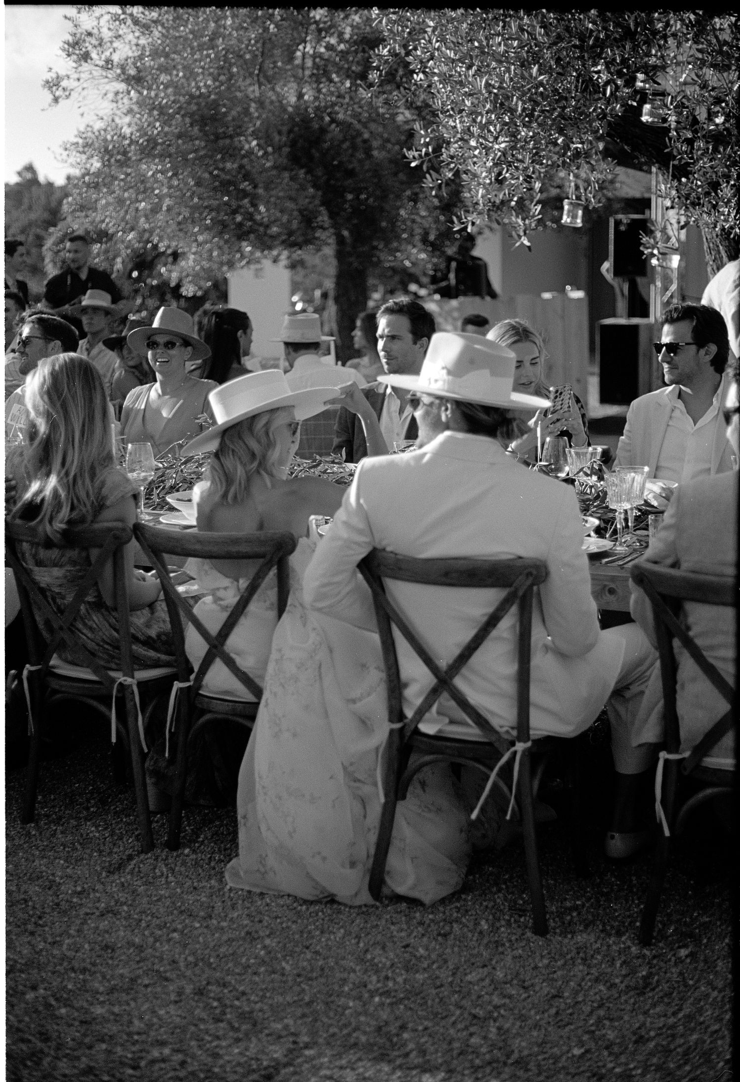 Black and white outdoor wedding reception with guests seated at tables under trees.