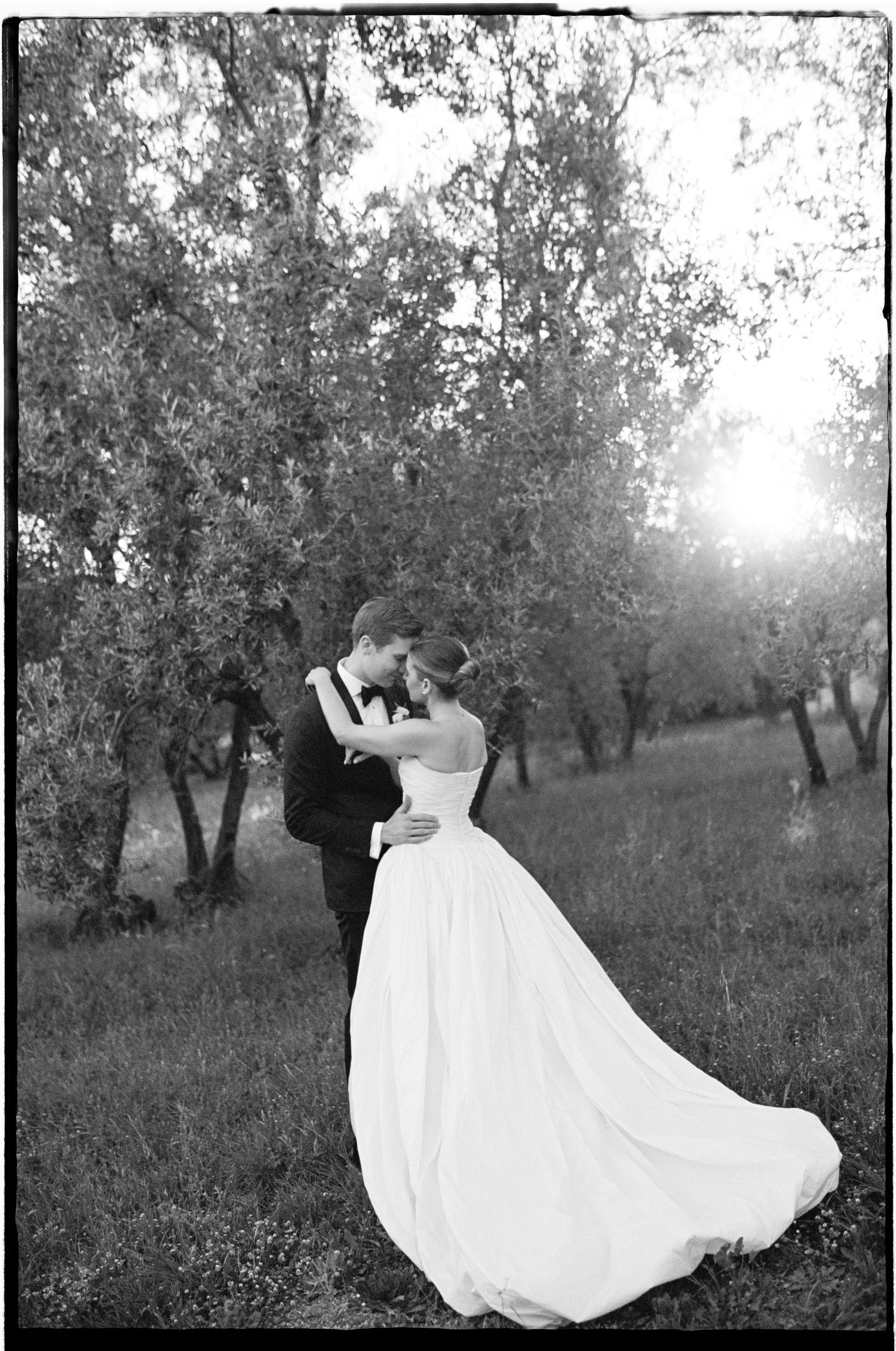 Black and white photo of a bride and groom embracing in a sunlit orchard, her gown flowing gracefully.