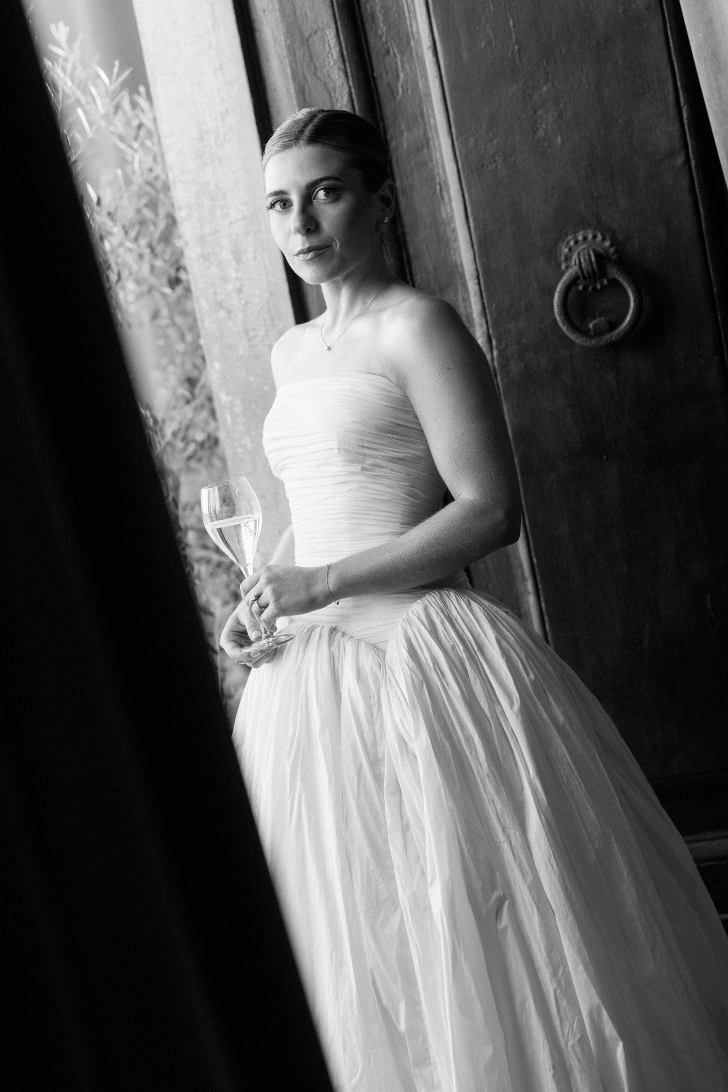 Elegant bride in strapless white gown holding champagne glass in black and white photo by ornate wooden door.