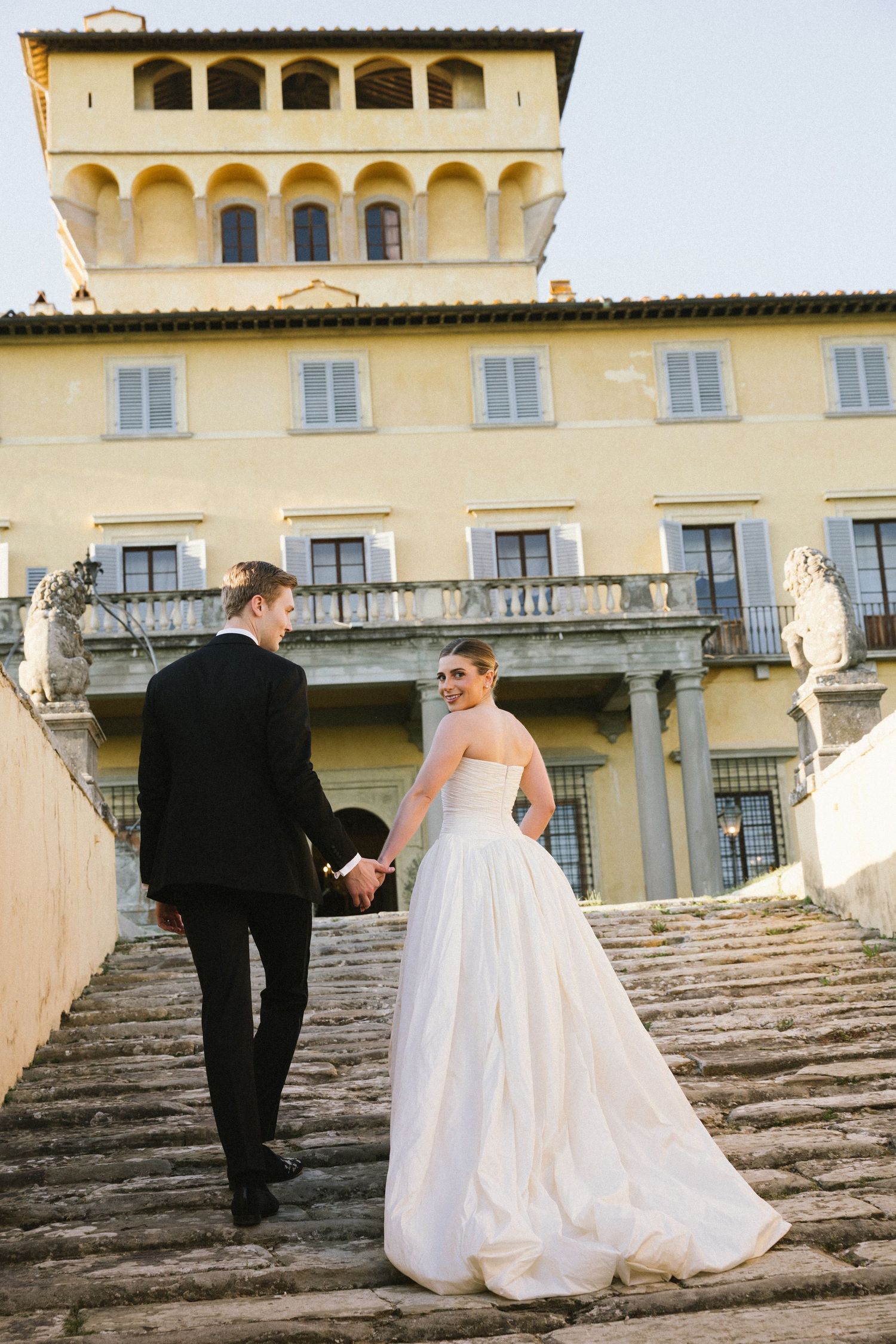 Bride and groom holding hands walking up stone steps of grand Italian Renaissance villa on wedding day.