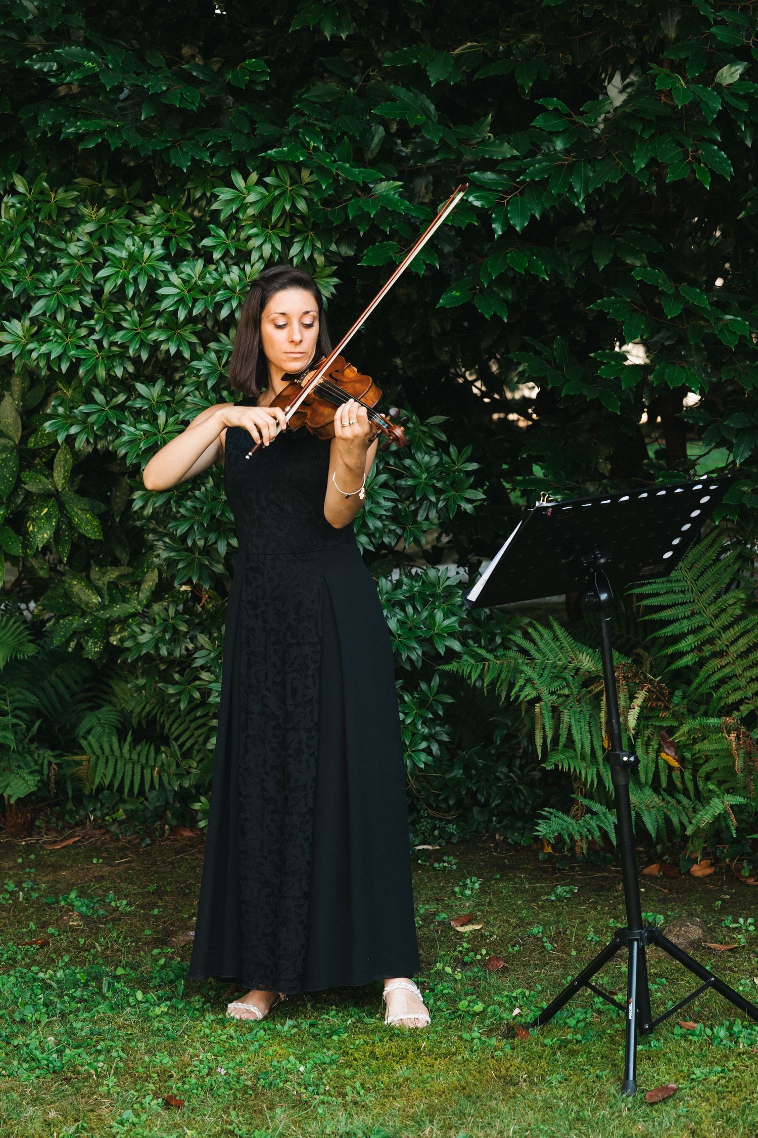 Woman in black dress playing violin outdoors with music stand, surrounded by lush green foliage.